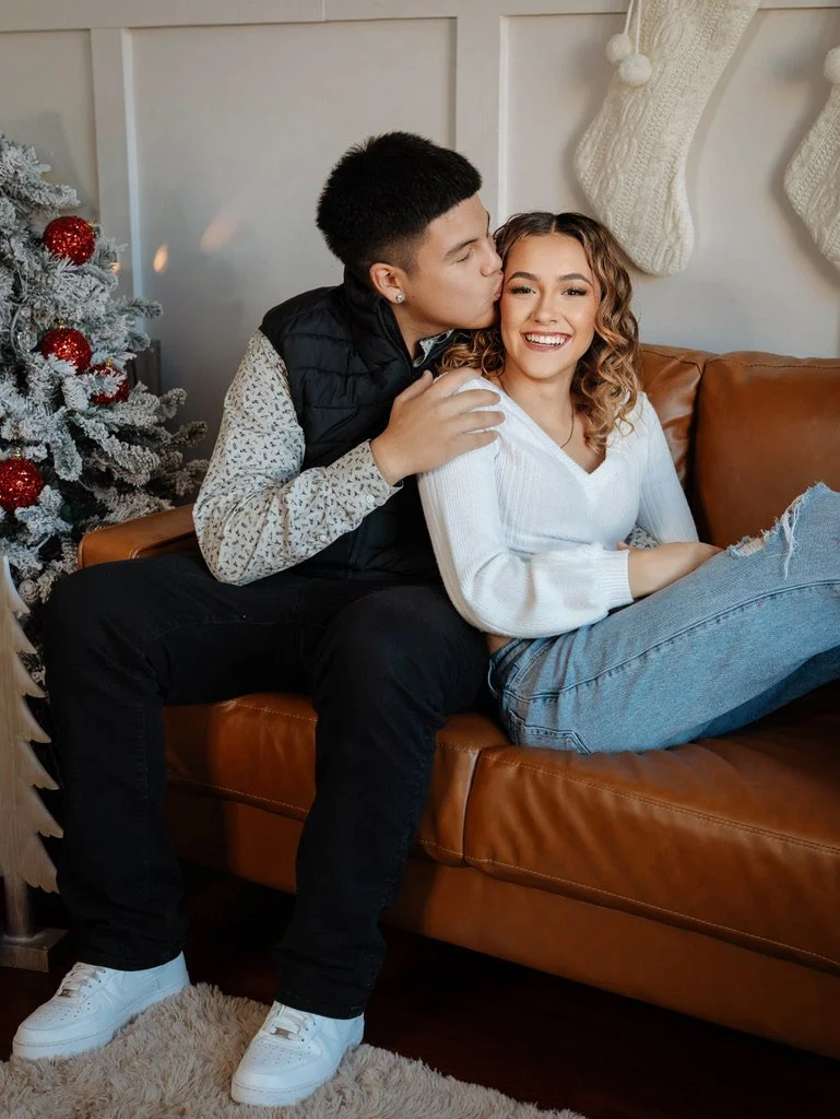 A young man is kissing a young woman on the cheek while sitting on a brown leather sofa. The woman is smiling and wearing a white sweater and light blue jeans. There is a decorated Christmas tree with red ornaments and two large Christmas stockings hanging on the wall behind them.