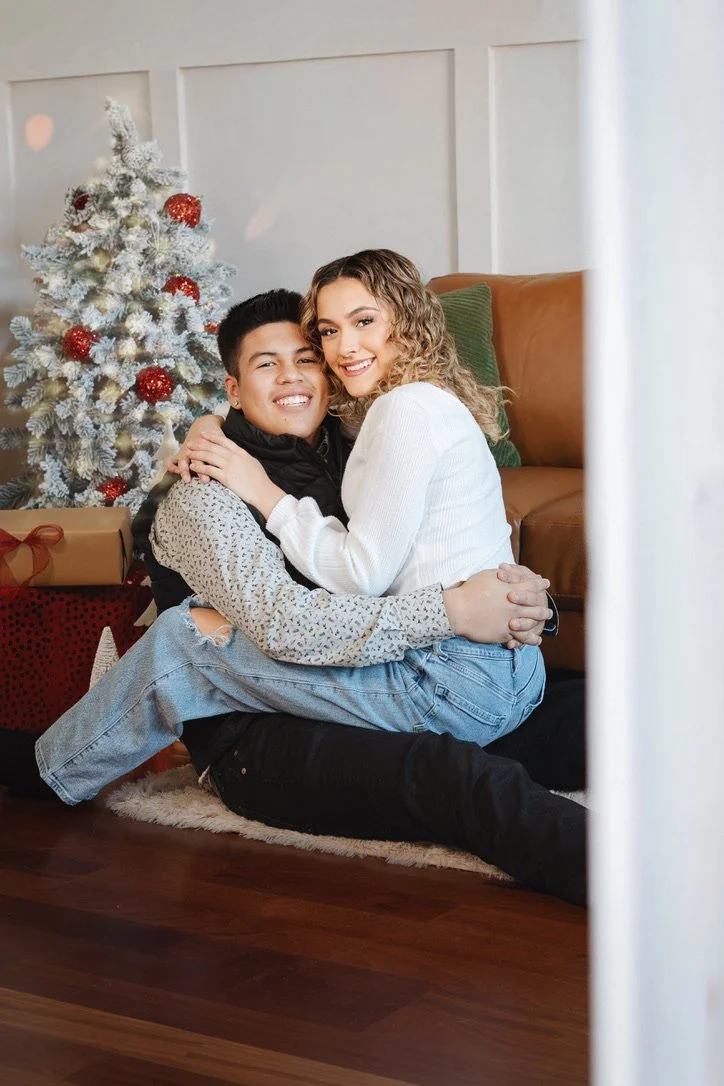 A young couple sitting on the floor in front of a Christmas tree, smiling and hugging each other indoors.