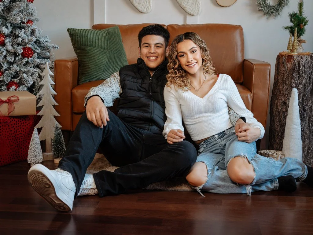A young man and woman sitting on the floor in front of a brown leather couch with Christmas decorations around them, including a Christmas tree, wrapped presents, and holiday ornaments, smiling for the camera.