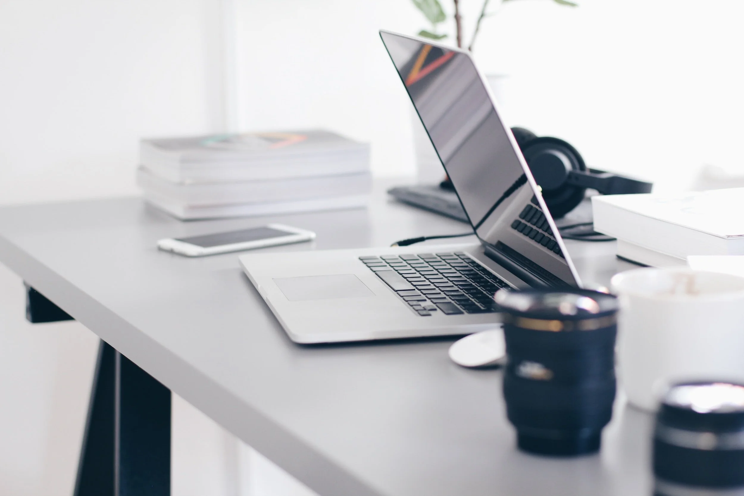 A workspace with a laptop, a smartphone, a pair of headphones, a stack of books, a coffee cup, and camera lenses on a white desk.