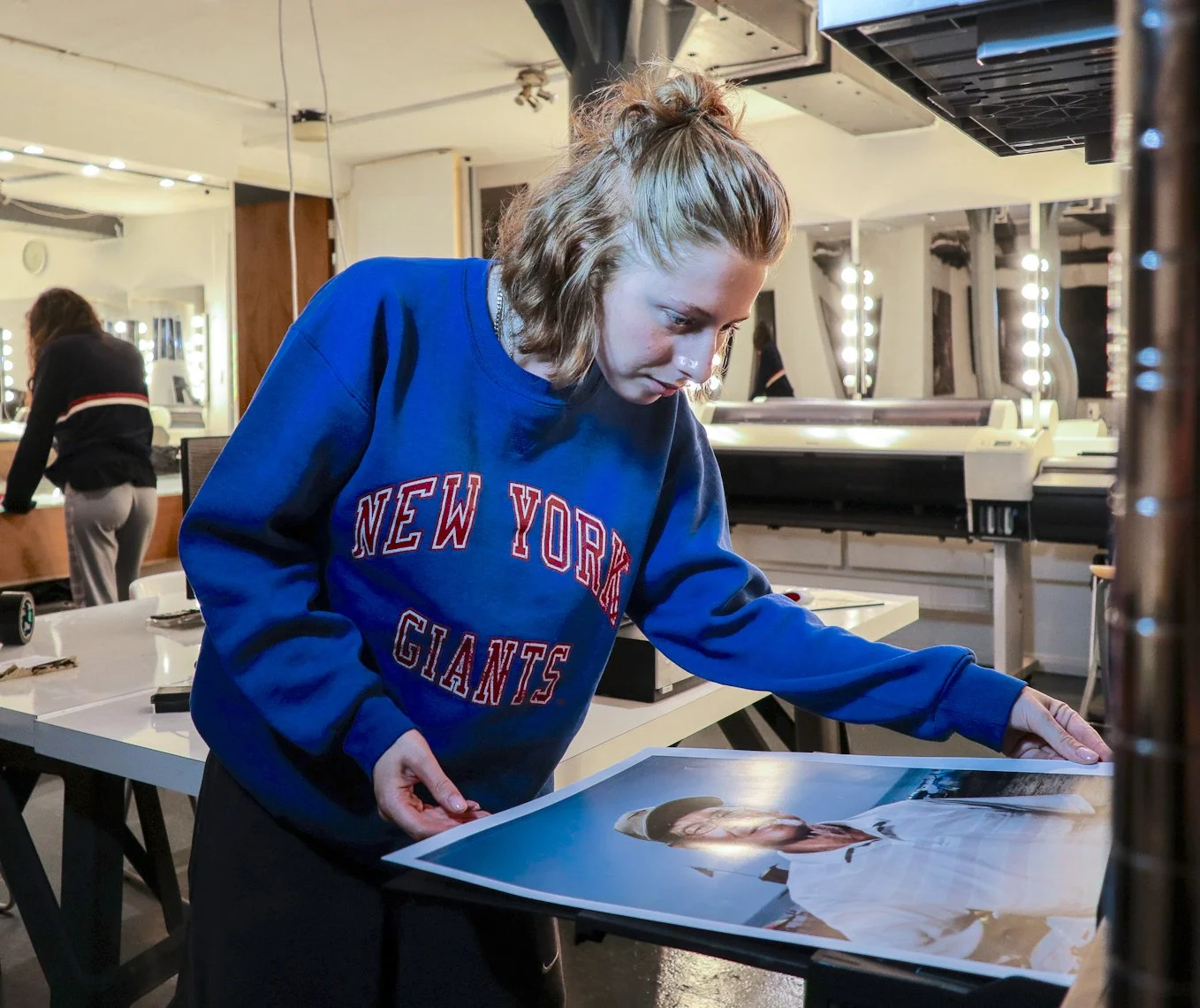 A woman with shoulder-length blonde hair wearing a blue sweatshirt with 'New York Giants' printed on it is examining a large photograph of a woman in a white outfit and hat, in a well-lit studio or salon with mirrors and lights on the walls in the background.