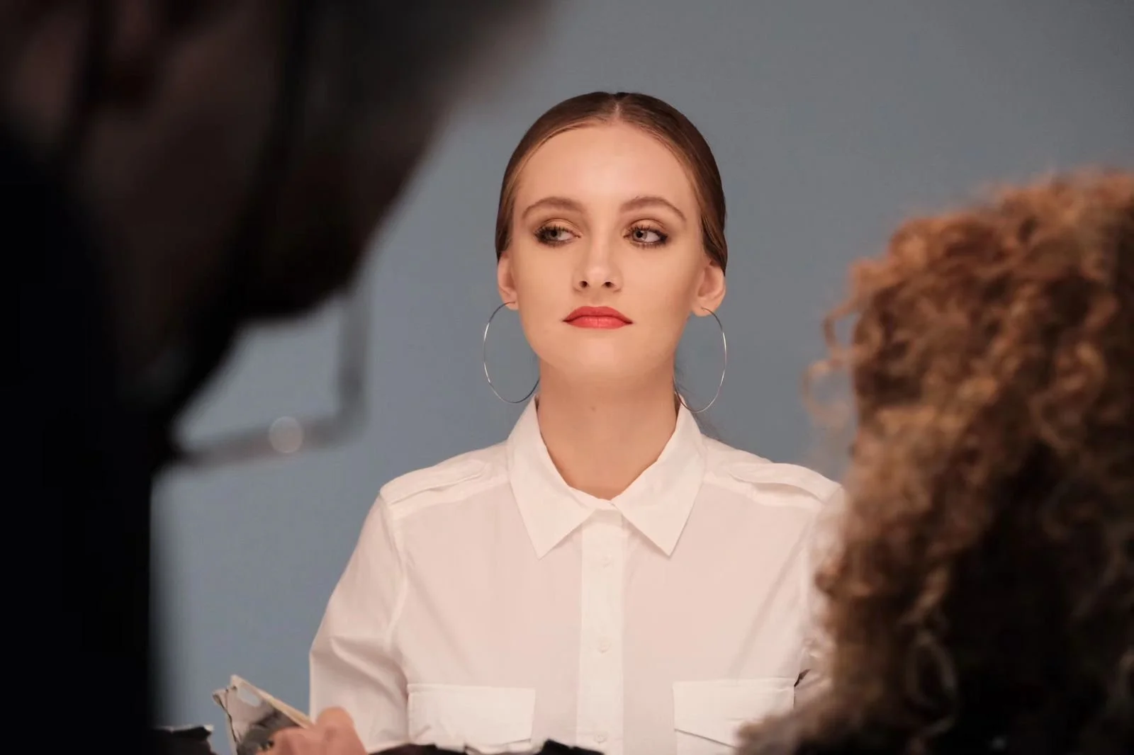 A young woman with light makeup, large hoop earrings, and a white collared shirt, looking to her left with a neutral expression, seated in front of a plain grey background.