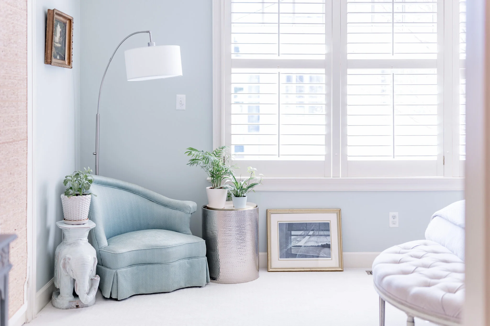 Bright living room corner with light blue walls, featuring a teal armchair and an elephant-shaped side table adorned with a potted plant. A metallic round table holds additional plants. A floor lamp and framed artwork are present, with white plantati