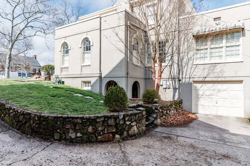 Exterior of a large, two-story house with arched windows and a garage. The house is surrounded by a stone retaining wall, manicured lawn, and leafless trees. A paved driveway leads to the garage.