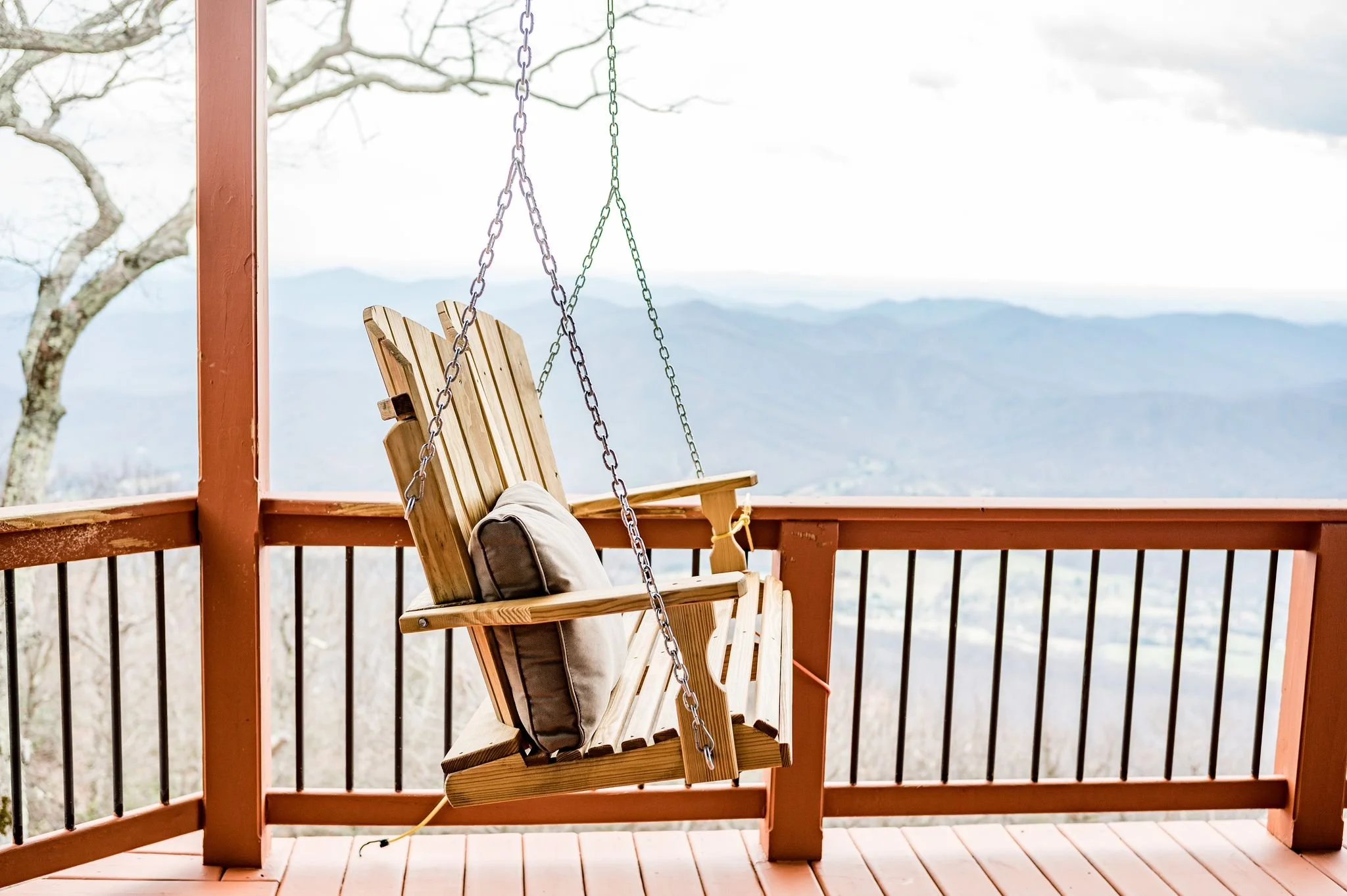Wooden porch swing with a cushion, overlooking a mountainous landscape under a cloudy sky.
