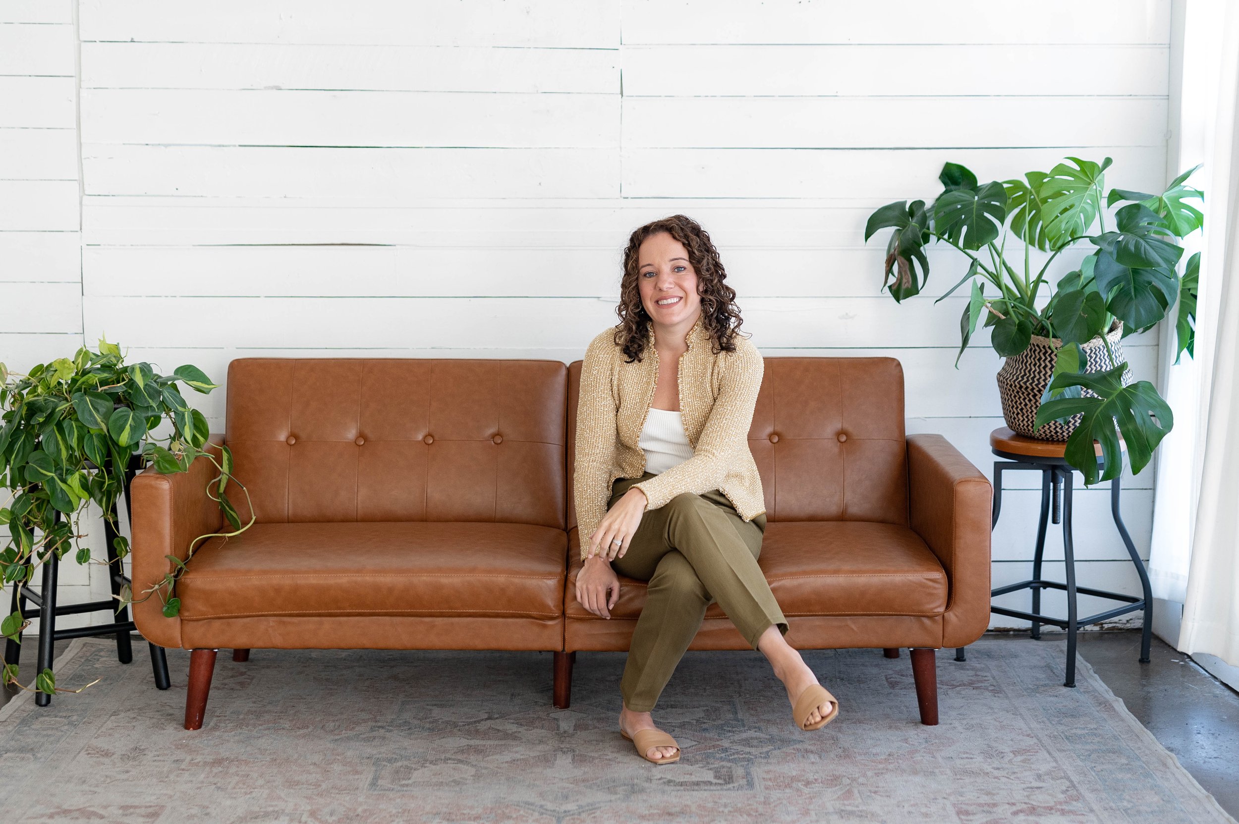 Woman sitting on a brown leather sofa, surrounded by houseplants, against a white wooden wall.