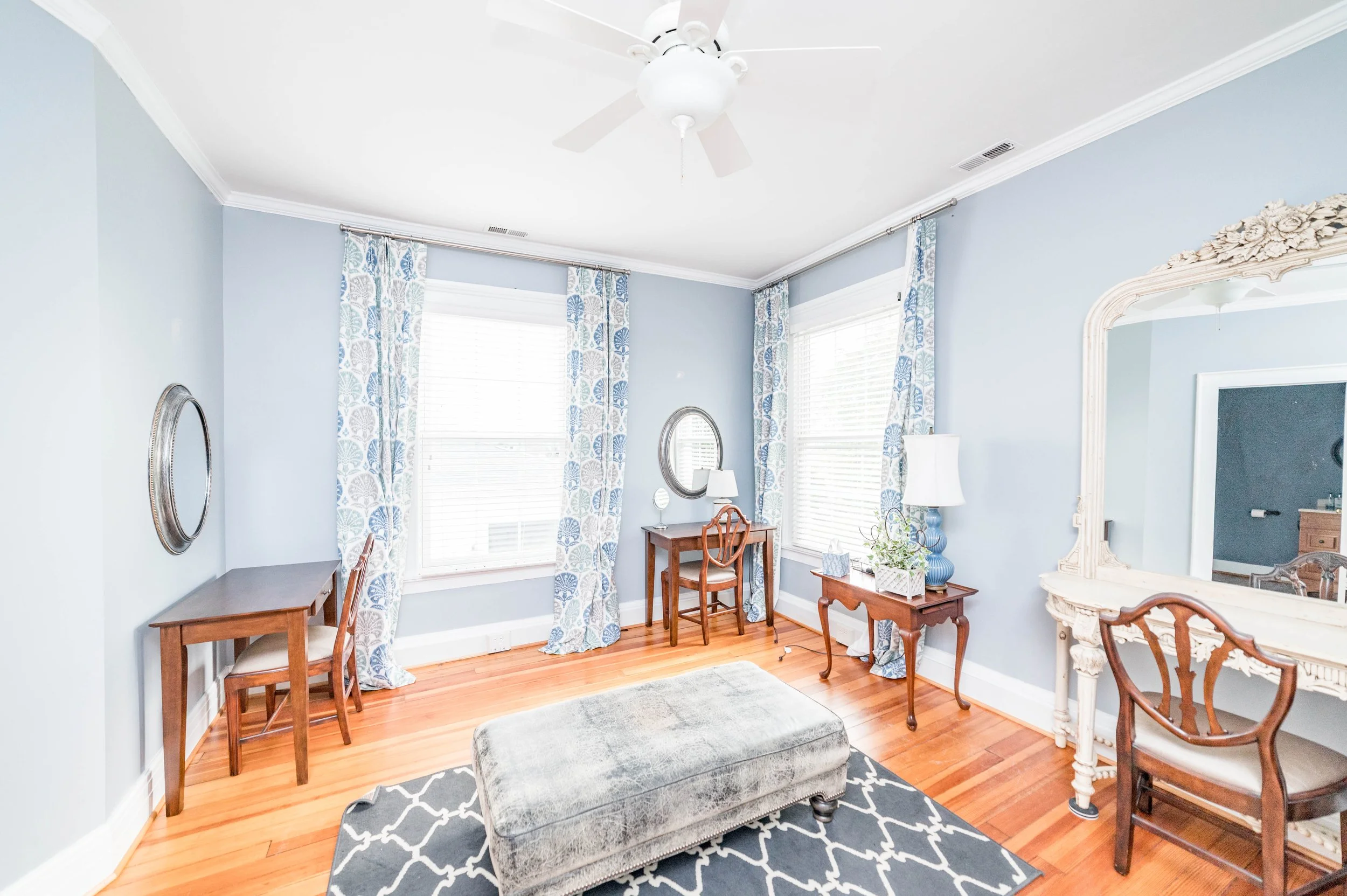 Cozy study room with blue walls, wooden desks and chairs, patterned curtains, a large mirror, and a central ottoman. There are decorative lamps on side tables and a ceiling fan.