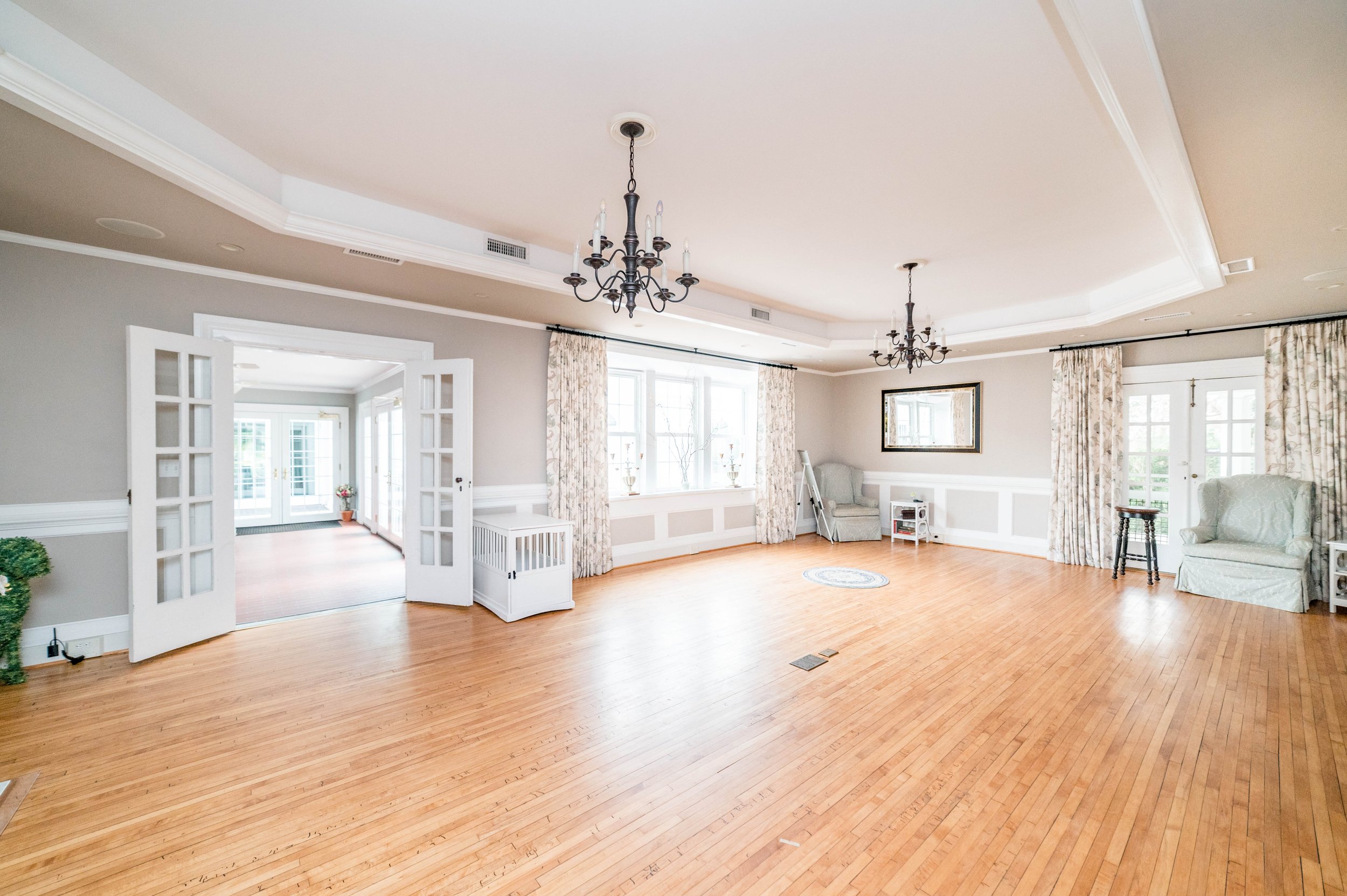 Spacious, bright living room with hardwood floors, white trim, and two chandeliers. Features large windows with floral curtains, a mirrored wall, armchairs, a small table, and French doors opening to another room.