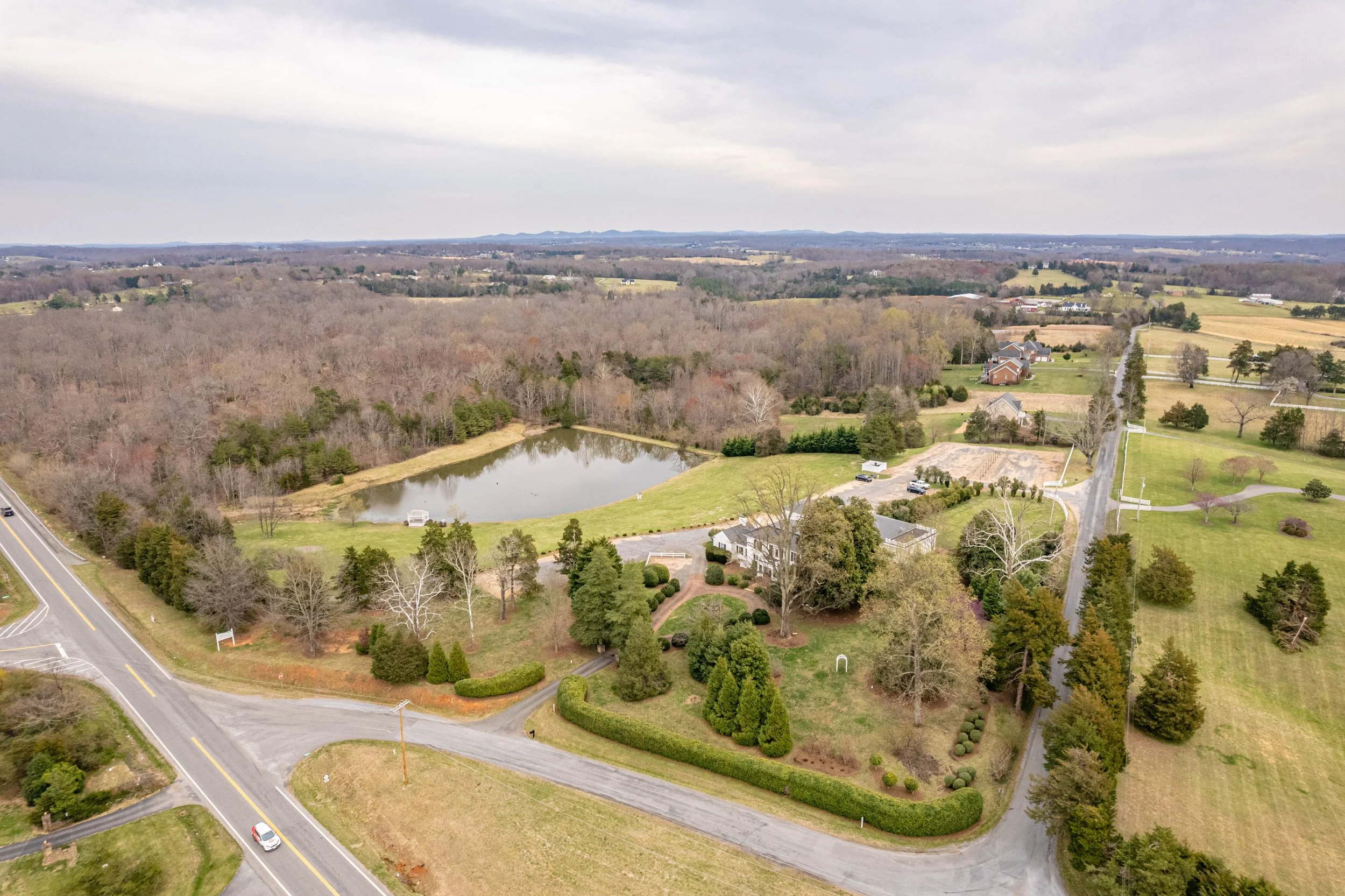 Aerial view of rural landscape with a pond, trees, roads, and scattered houses on grassy fields.