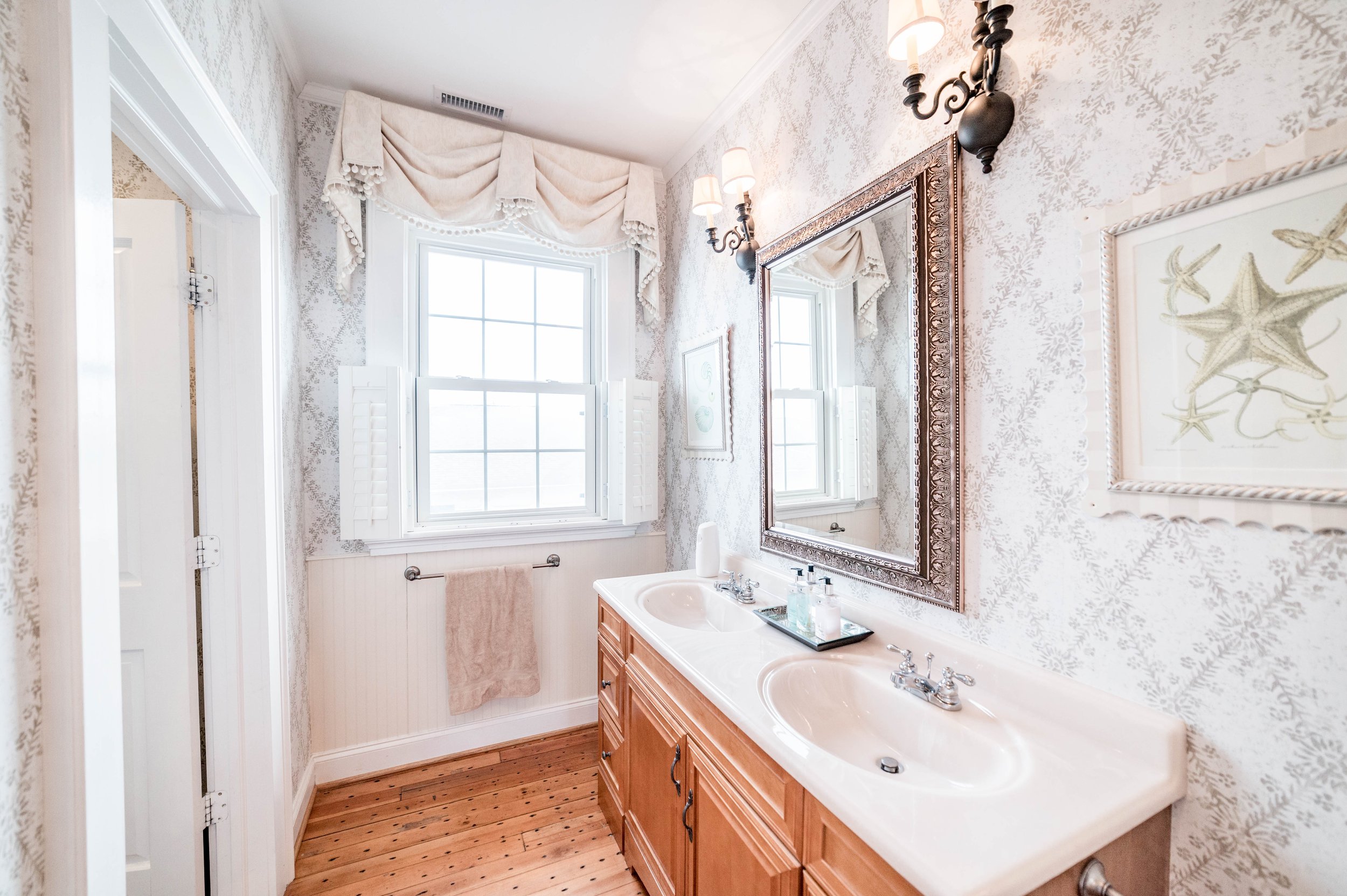 Elegant bathroom with double sink vanity, ornate mirror, wall sconces, starfish artwork, large window with draped curtains, and hardwood floor.