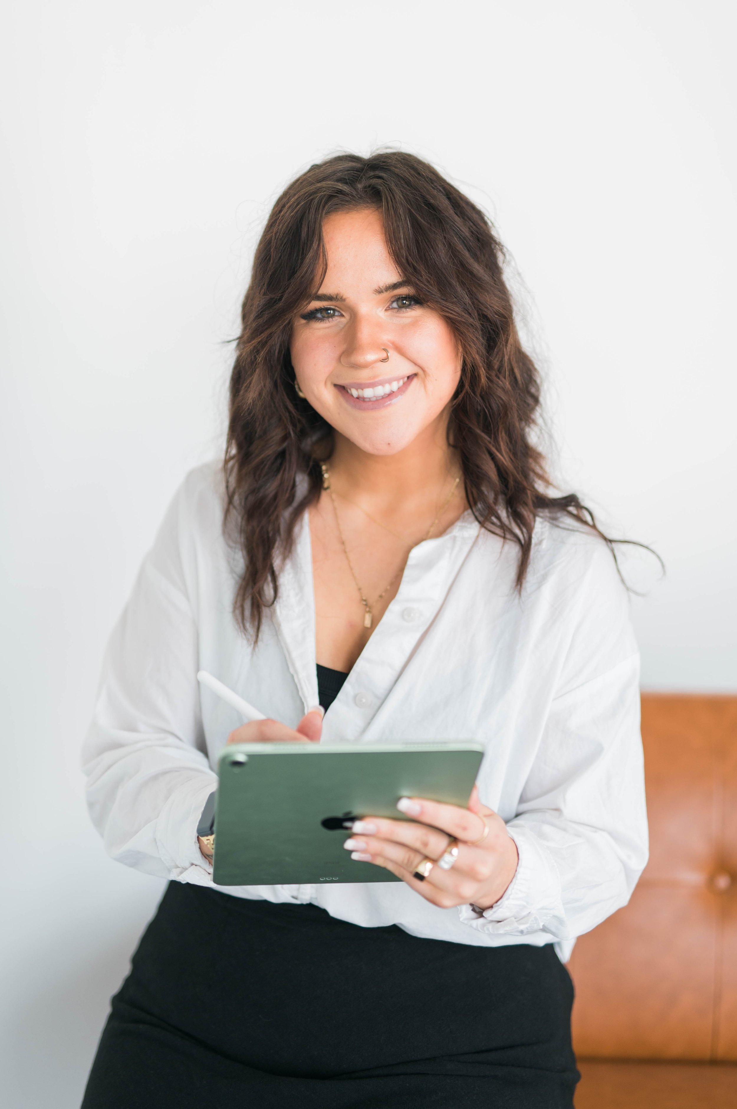 A woman with long dark hair is smiling while holding a tablet and stylus. She is wearing a white button-up shirt and a black skirt, sitting against a white background.