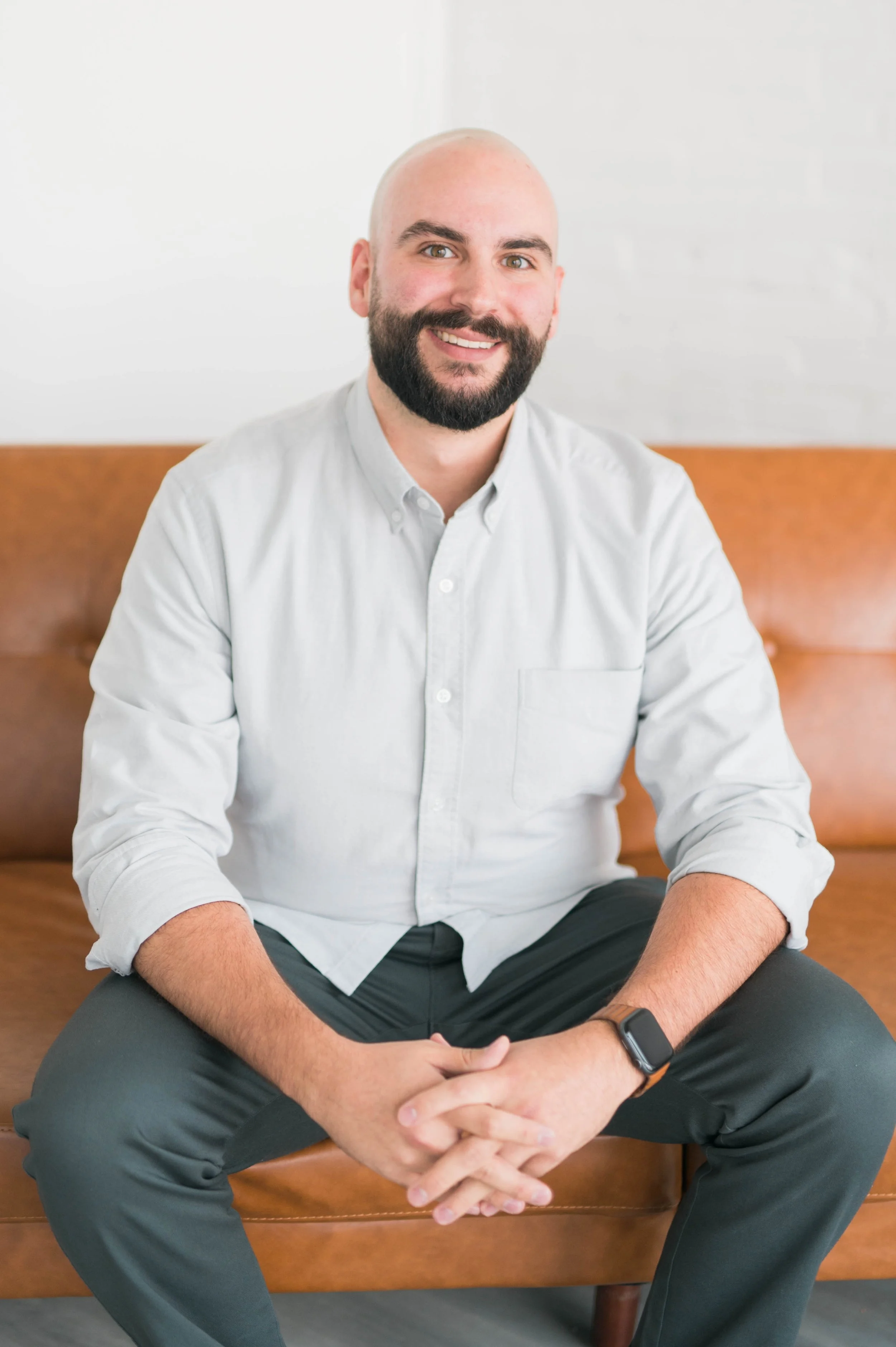 Bald man with a beard sitting on a brown leather couch, wearing a light gray button-up shirt and black pants, smiling with hands clasped.