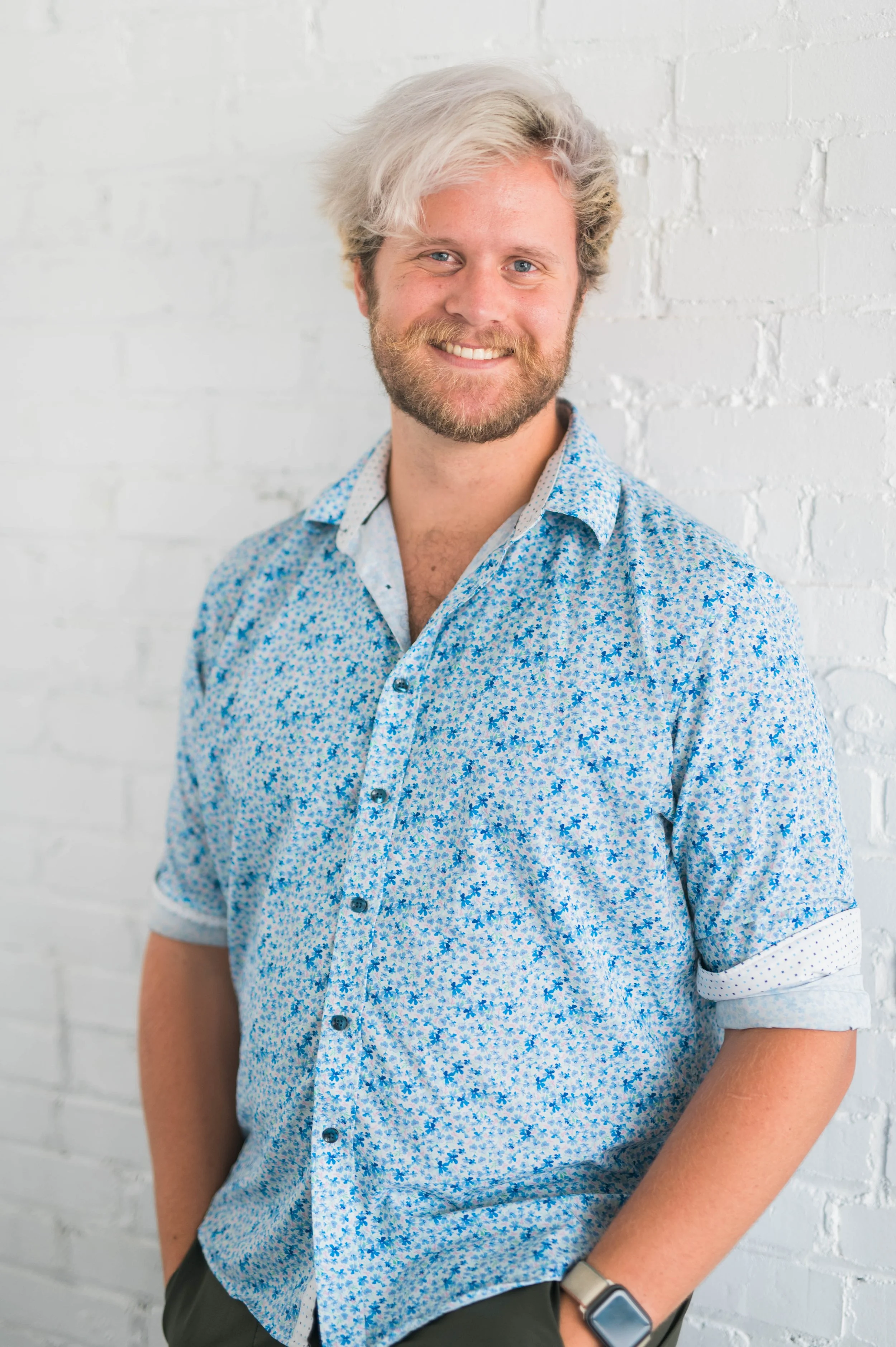 Smiling person with light hair wearing a blue floral shirt standing against a white brick wall.