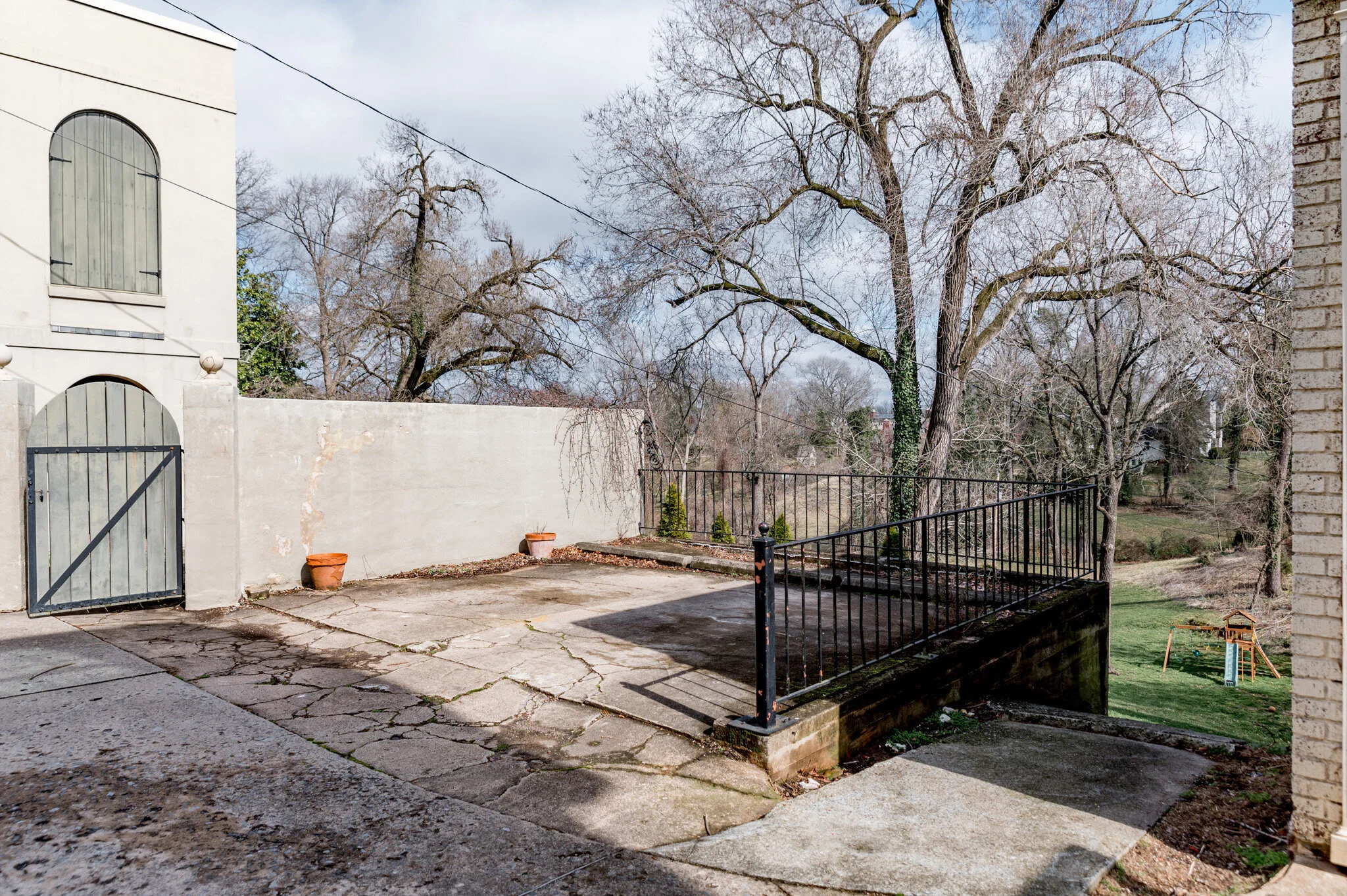 Outdoor area with a concrete patio, black metal railing, potted plants against a stucco wall, and leafless trees. In the background, a grassy field and a children's playset are visible.
