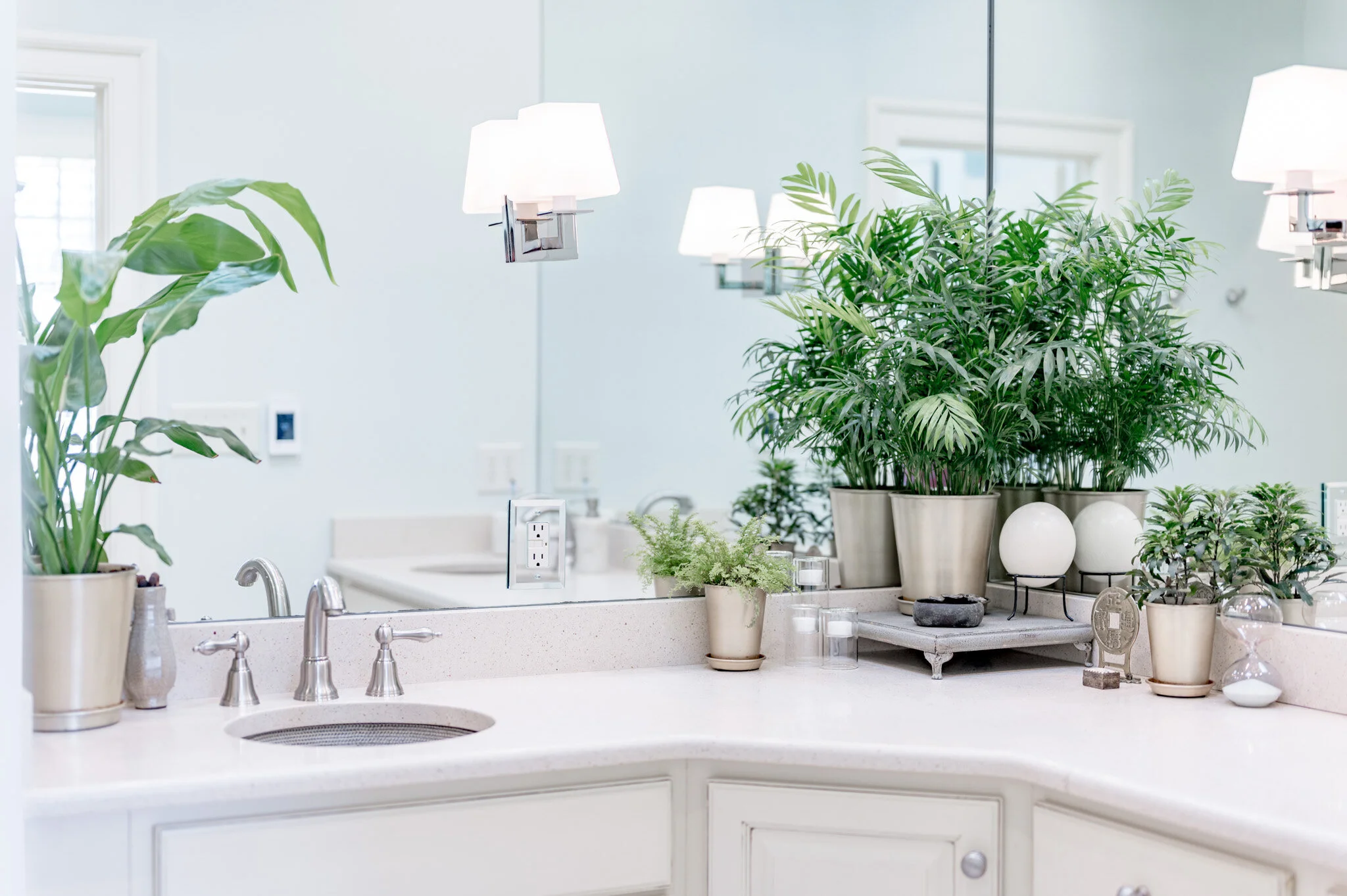 Bathroom vanity with potted plants, round light fixture, silver faucet, and mirror reflecting the decor.
