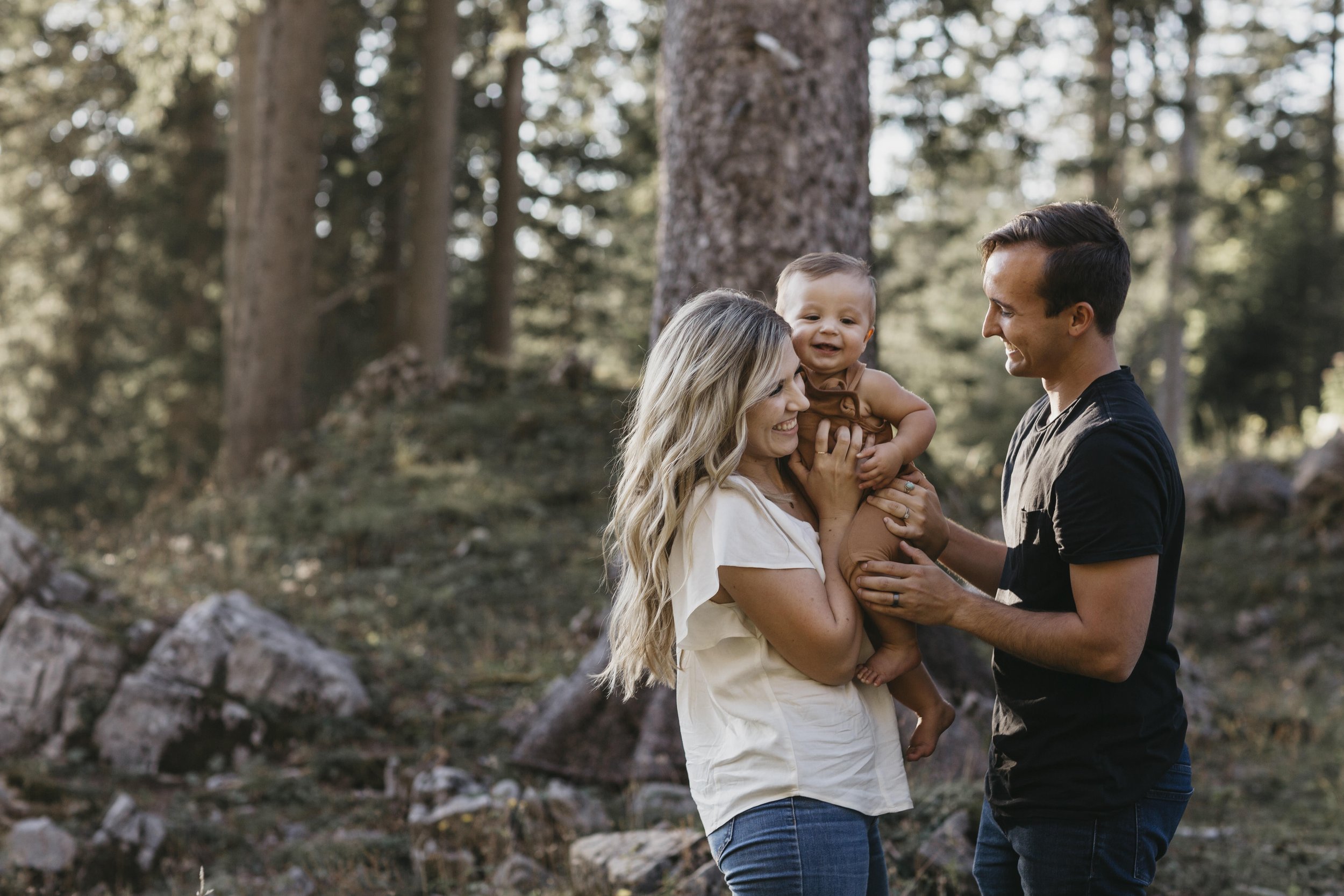 A smiling couple stands in a wooded area, with the woman holding a happy baby in her arms. The man stands beside them, also smiling, and they are surrounded by trees and rocks.