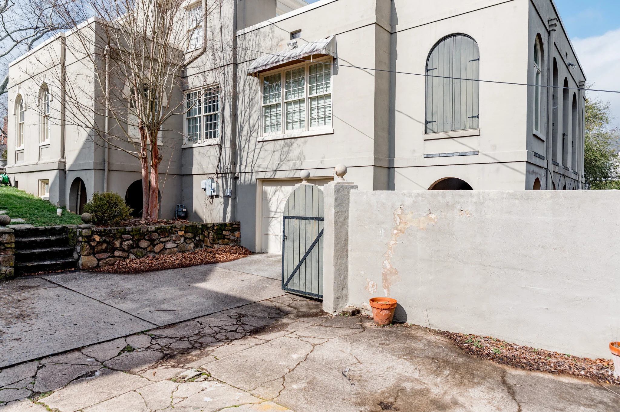 Exterior view of a building with arched windows and a garage. The building has a light gray facade and a gated driveway with a cracked stone surface. There is a stone retaining wall on the left with steps leading to a grassy area. A leafless tree and