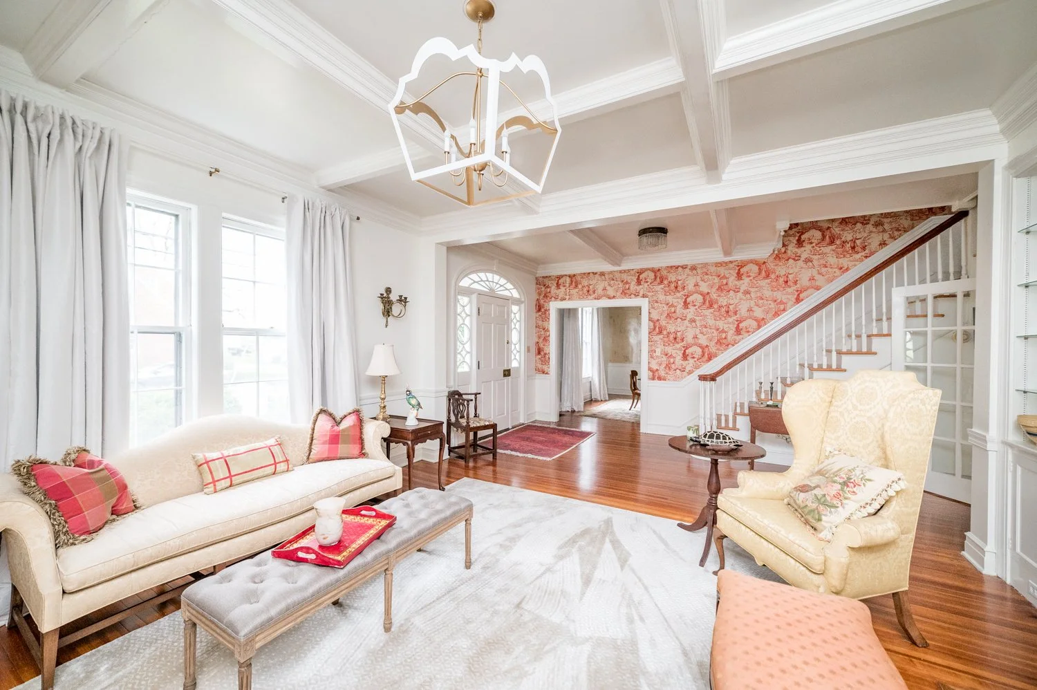 Elegant living room with white walls, coffered ceiling, large windows with curtains, cream sofa with red pillows, floral armchair, patterned wallpaper, hardwood floors, and chandelier.