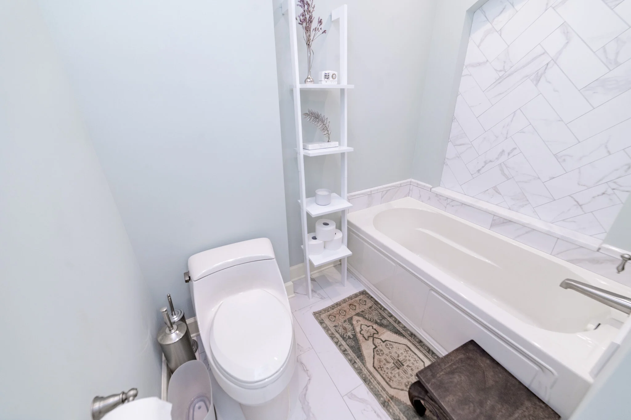 Bathroom with white toilet, bathtub, herringbone tile, ladder shelf with decor and toilet paper, and a patterned rug.