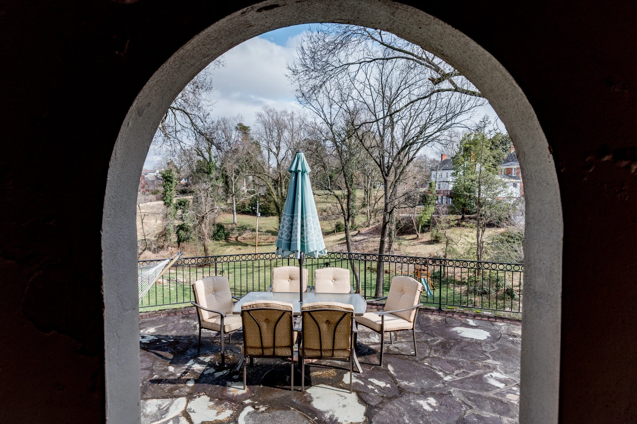 Outdoor patio with table, chairs, and closed umbrella, viewed through an arched doorway, with trees and a house in the background.