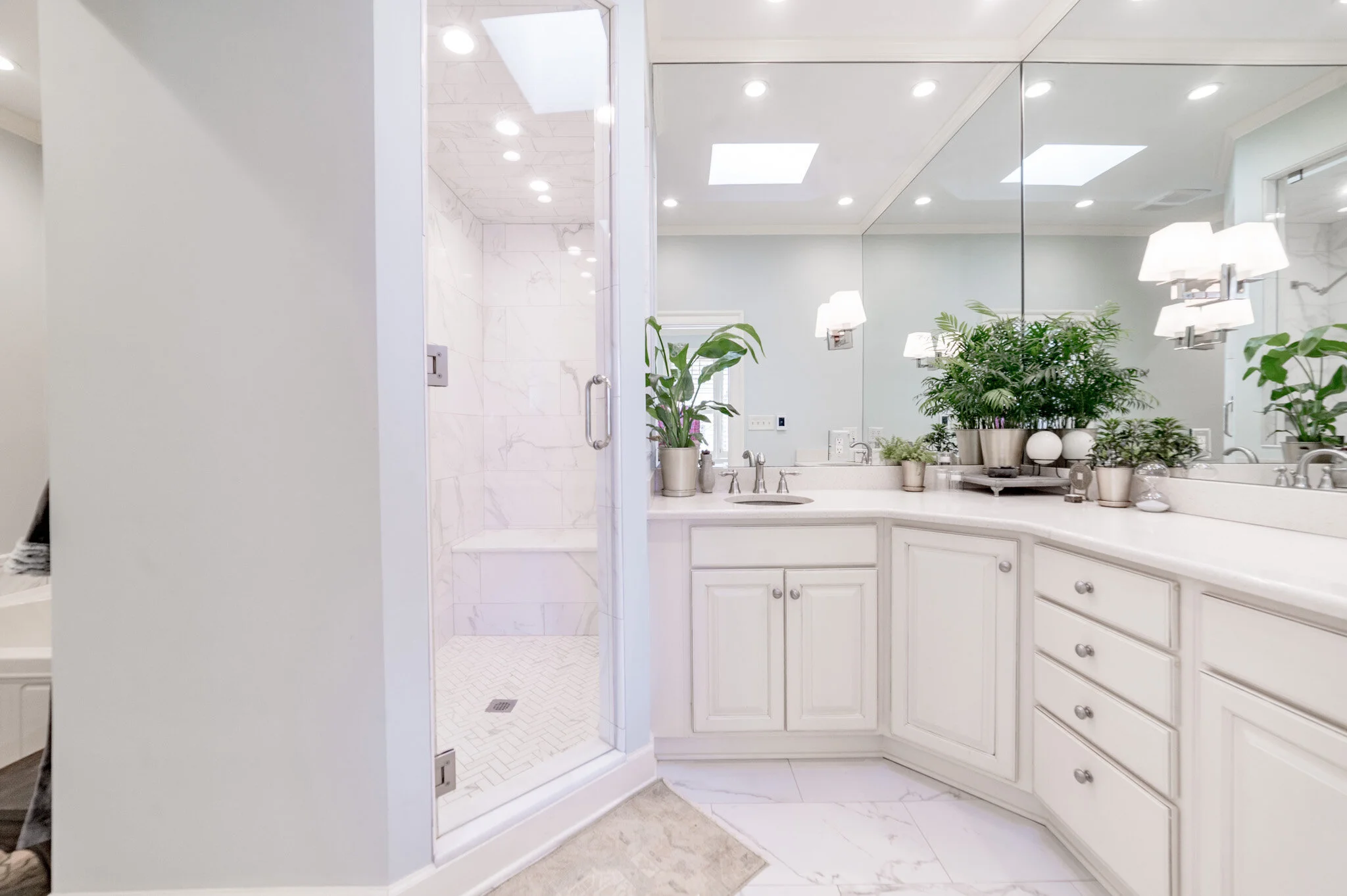 Stylish modern bathroom with white cabinets, large mirror, potted plants, and glass-enclosed shower with marble tile.
