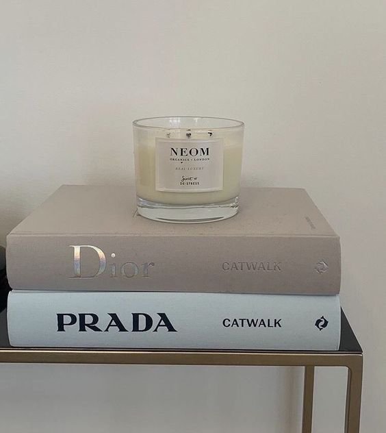 A candle labeled 'NEOM' sits on top of two fashion books titled 'Dior' and 'Prada' on a shelf.