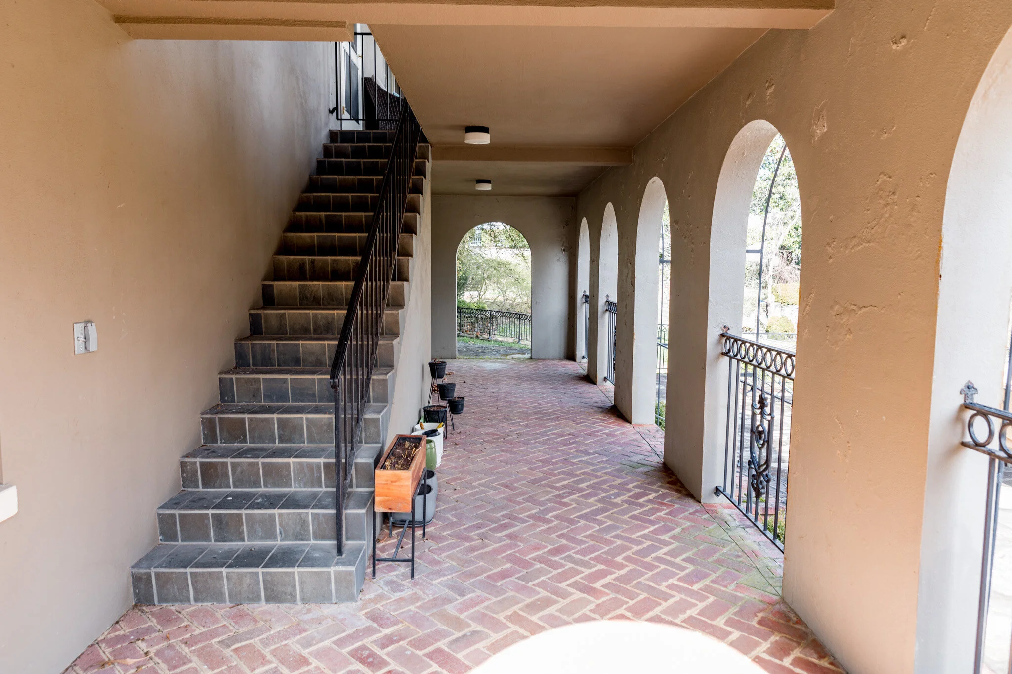 Outdoor corridor with brick flooring, arched windows, and a staircase on the left. The corridor has a beige wall and ceiling with light fixtures. Plants and pots line the pathway.