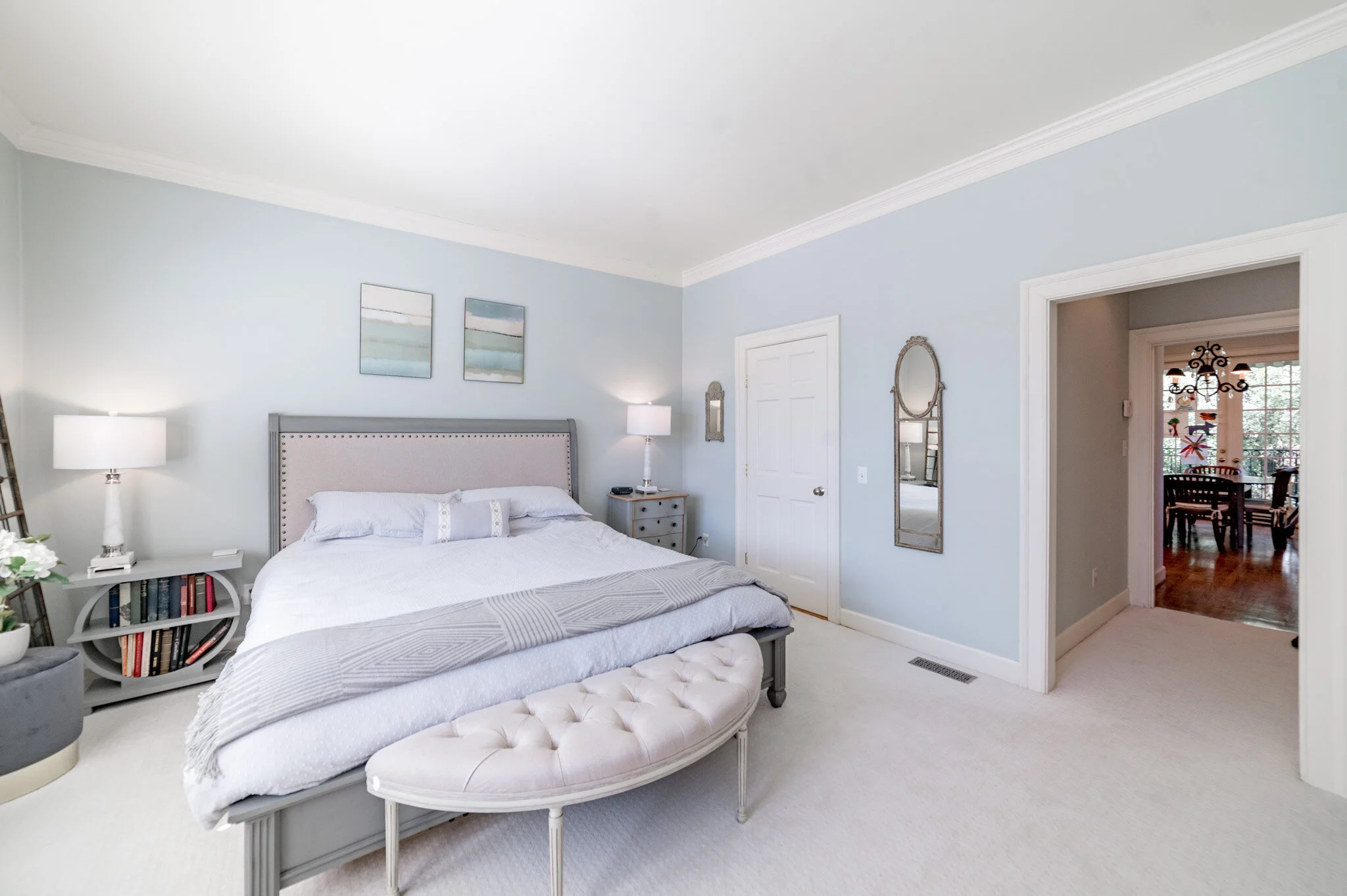 Interior of a modern bedroom with a large bed, light blue walls, and a decorative bench. Two abstract artworks hang above the bed, and there are matching nightstands with lamps on each side. A round mirror is mounted on the wall, and an open door rev