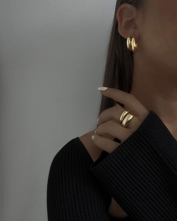 Woman wearing gold hoop earrings and a gold ring, black textured sweater, and white nail polish.
