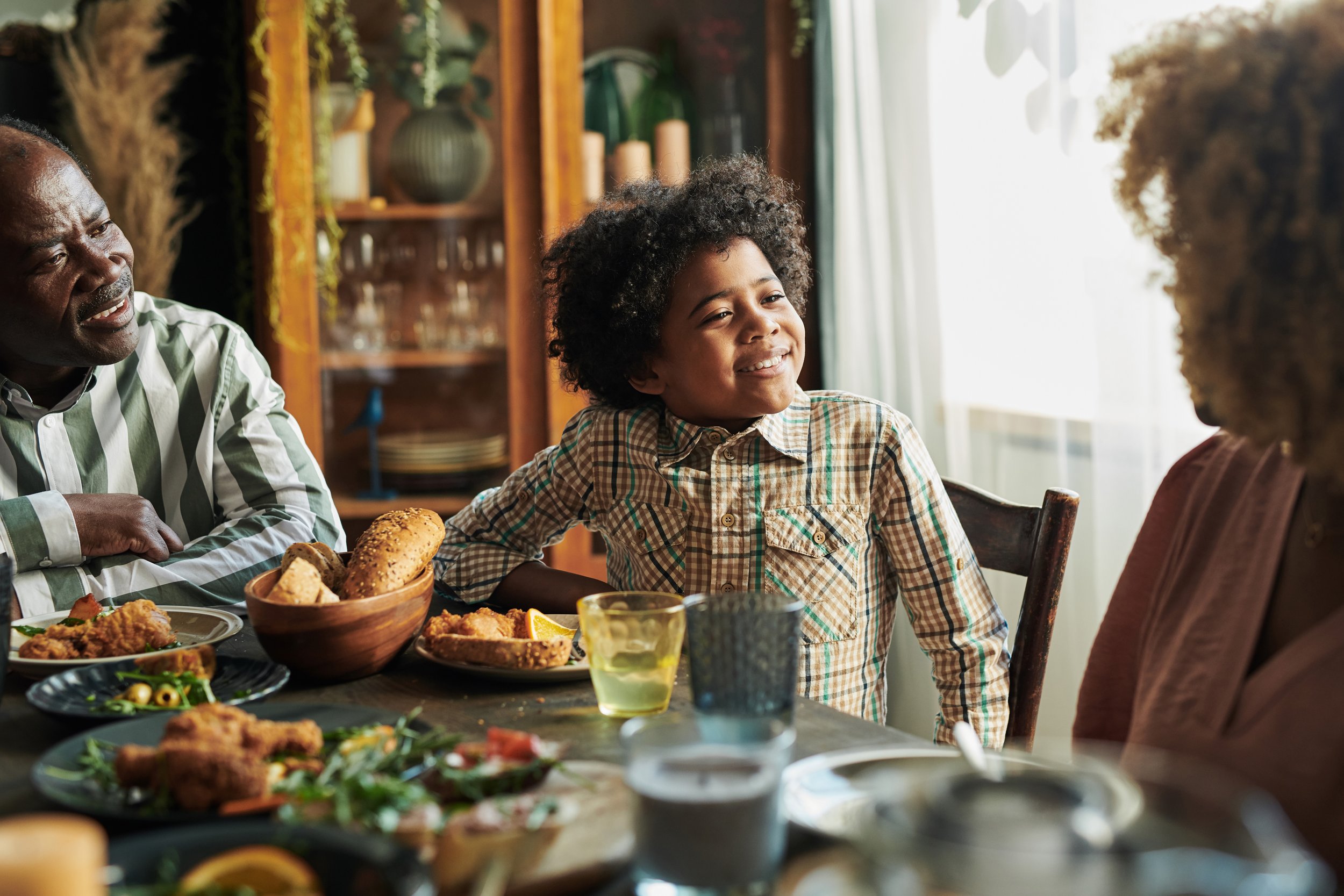 A family having a meal together, with a focus on a smiling child in a plaid shirt seated at a table with food, including bread and fried chicken. Other family members are seated nearby, engaging in conversation.
