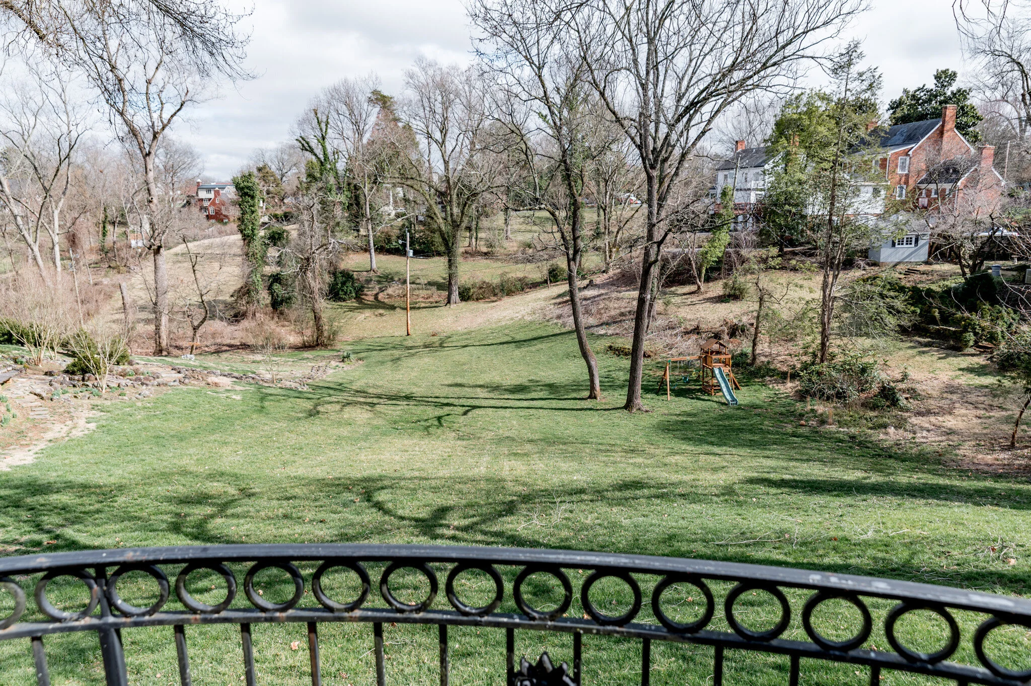 View of a large grassy backyard with trees, a metal railing in the foreground, a wooden swing set, and houses in the background.