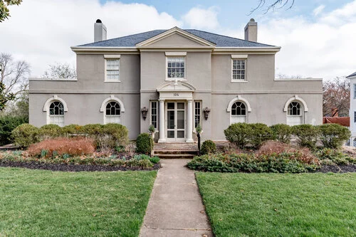 Front view of a large, classic-style house with a symmetrical design, featuring a central entrance with columns, multiple windows, and surrounded by landscaped shrubs and a lawn.