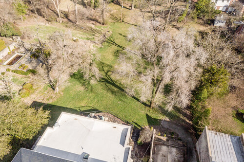 Aerial view of a residential area with a large grassy yard surrounded by trees, a white-roofed building, and a paved driveway.