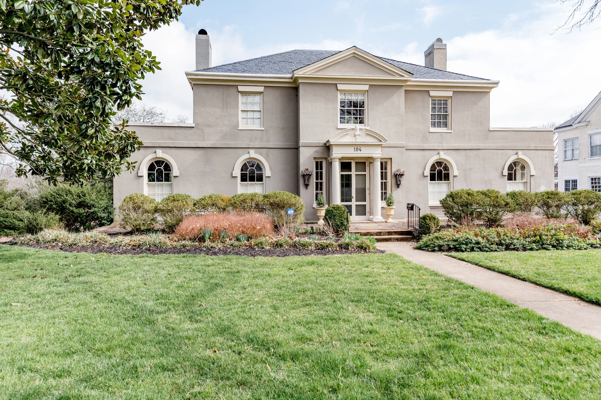 Large two-story house with beige stucco exterior, arched windows, and lawn. Features symmetrical landscaping, a pathway leading to the entrance, and tall chimneys. Detached neighboring house visible.
