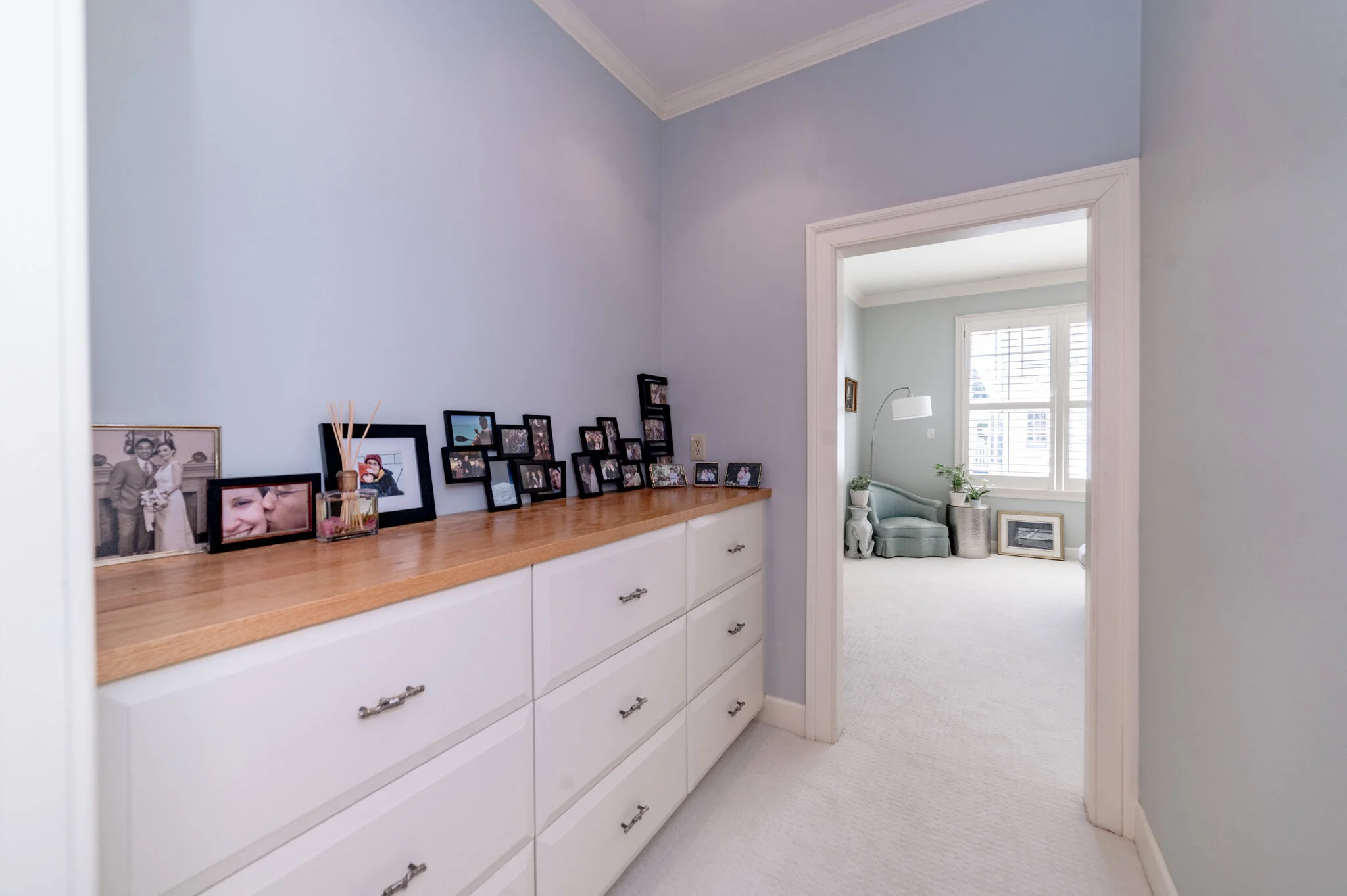 Hallway with white drawers featuring framed photos, leading to a room with a light blue armchair, a floor lamp, and large windows with white shutters.