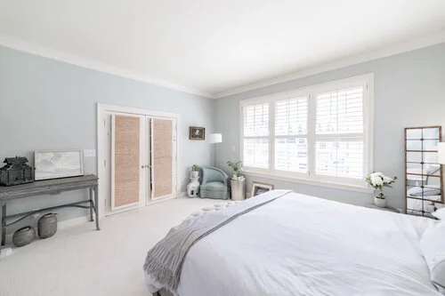 Bright bedroom with a neatly made bed, light blue walls, large window with shutters, a chair, and decorative items. Room features white bedding, and wicker basket decor. A standing mirror and small desk are present.