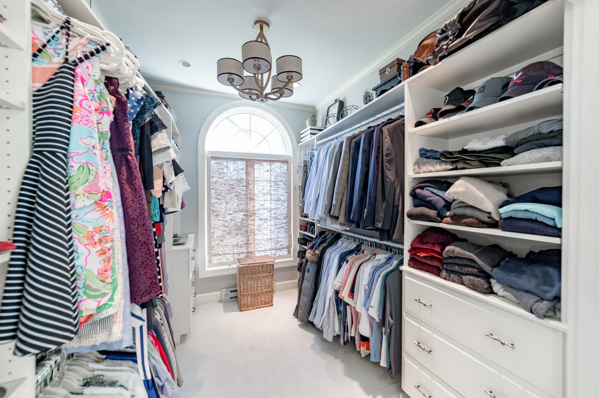 Walk-in closet with organized clothing, suits, shirts, dresses, and folded sweaters on shelves, beneath a hanging light fixture; a window with blinds at the far end.