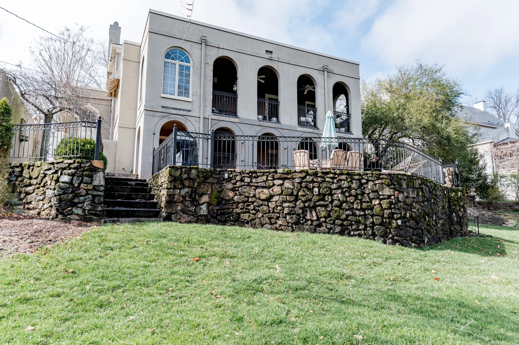 Two-story stone house with arched windows and balconies, surrounded by a stone terrace and iron railings, set on a grassy lawn with a staircase leading up.