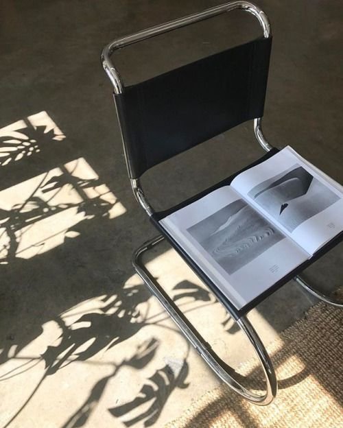 Metal chair with black seat holding an open art book on a concrete floor, alongside shadows of plant leaves.