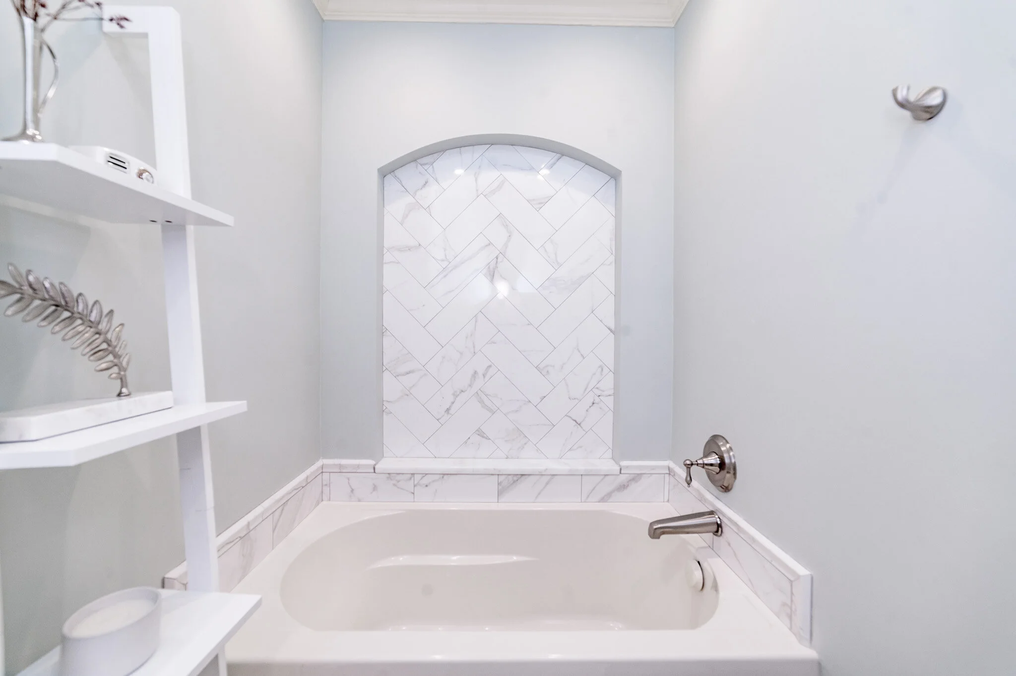 Bathroom with a white bathtub and marble tile backsplash, gray walls, and white shelving.