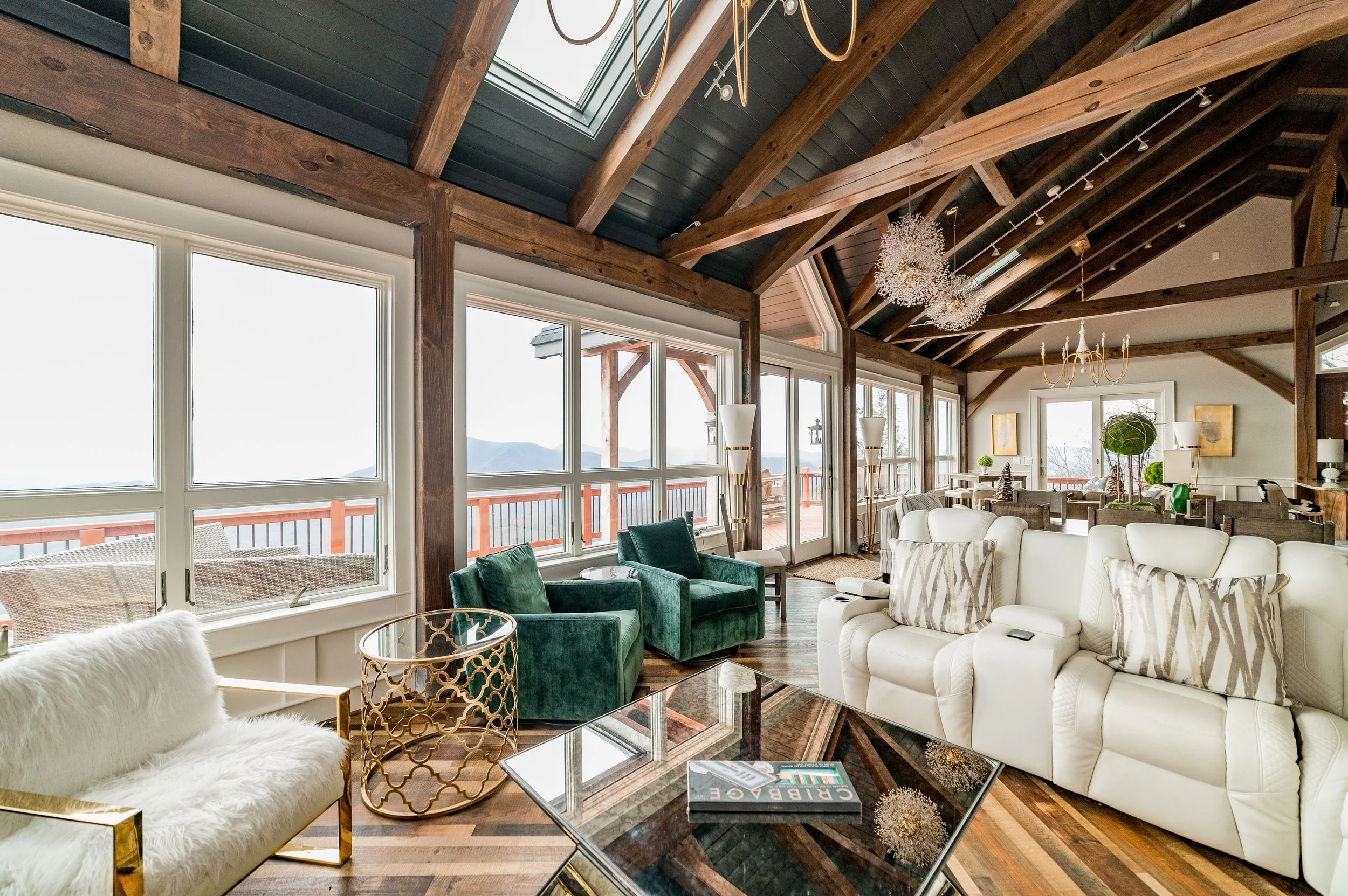 Luxurious living room with large windows, wooden beams, modern furniture, green velvet chairs, a fluffy white chair, glass coffee tables, and decorative chandeliers, offering a panoramic mountain view.