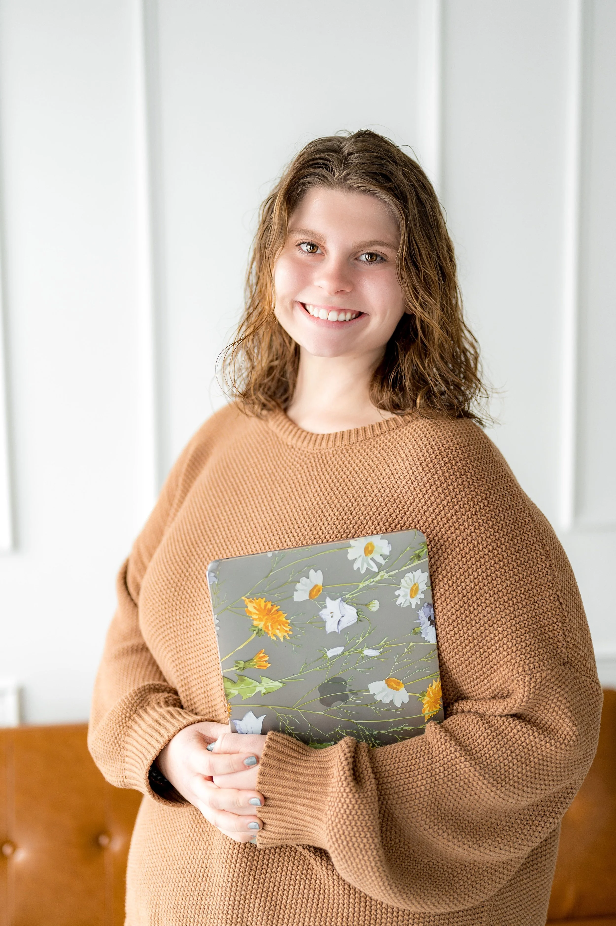 Smiling person in brown sweater holding a floral-patterned laptop standing in front of a white wall.