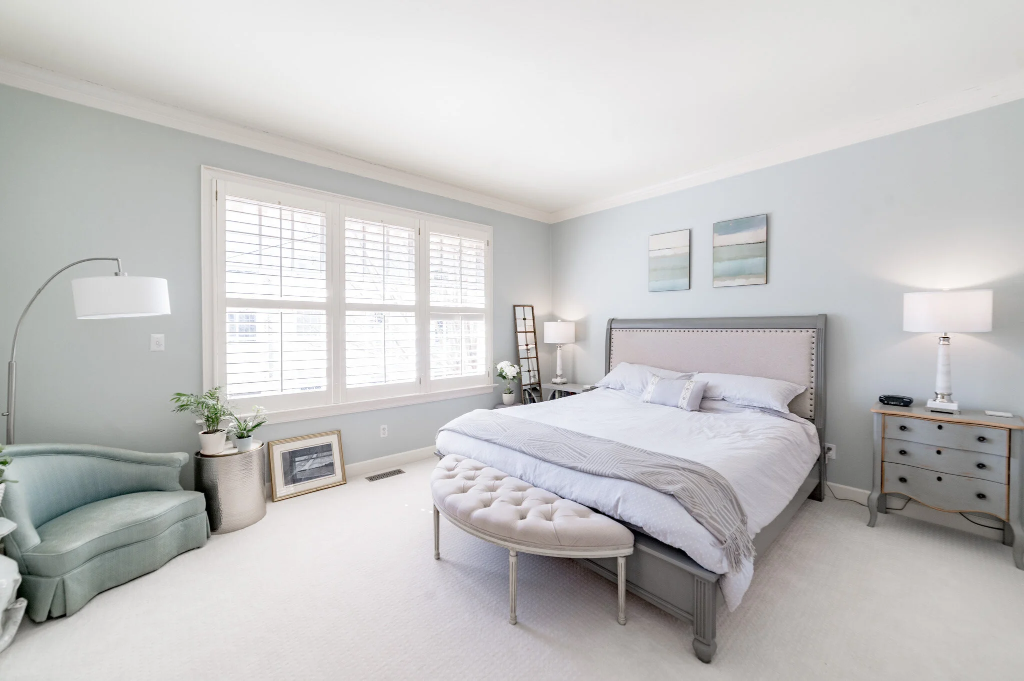 Elegant bedroom with a king-sized bed, light blue walls, and two bedside lamps. A tufted bench sits at the foot of the bed, and a plush chair is near a window with white shutters. Decorative plants and framed pictures complete the decor.