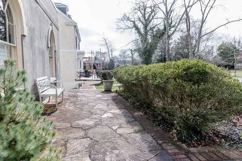 Stone patio with benches and potted plants next to a building, alongside hedges and bare trees.
