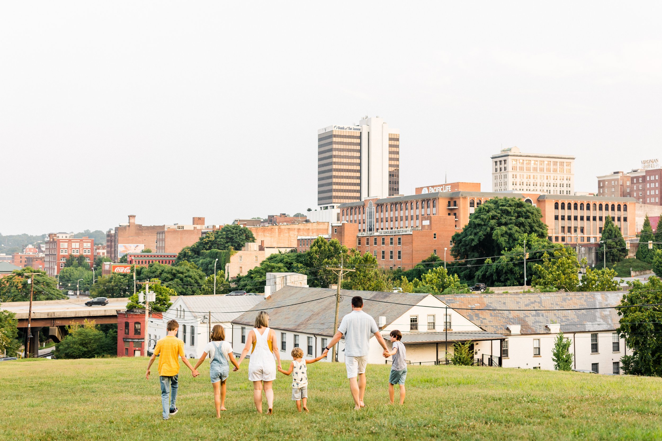 A family of six walking hand-in-hand on a grassy field with a cityscape in the background, including buildings and a bridge.