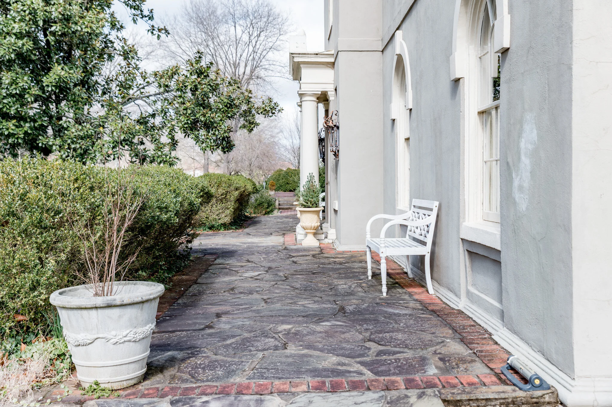 Outdoor stone patio with white bench, planters, and green shrubs along walkway, adjacent to gray building with arched windows
