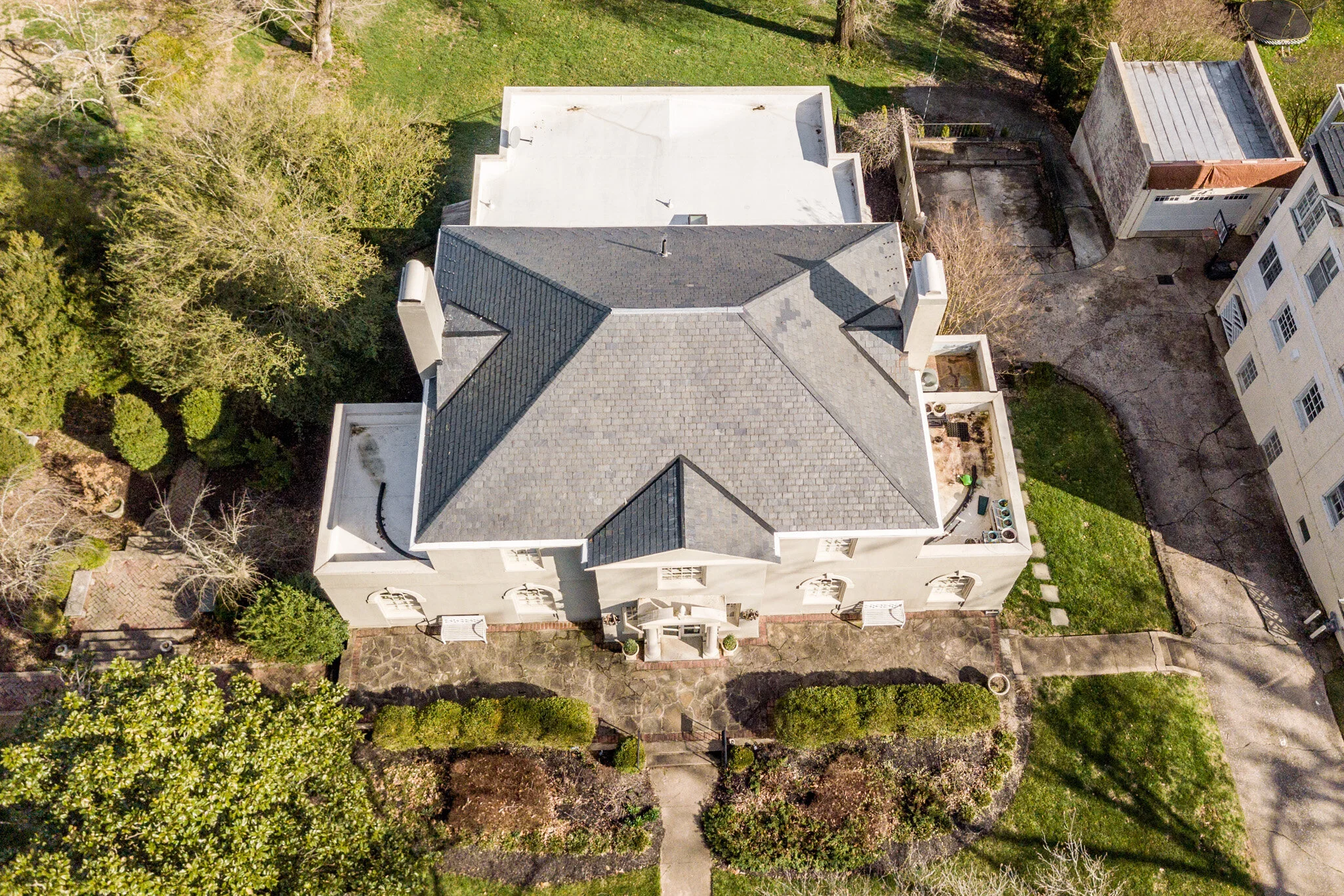 Aerial view of a large residential house with a gray tiled roof, surrounded by landscaped gardens and greenery, including a paved driveway and a small courtyard.
