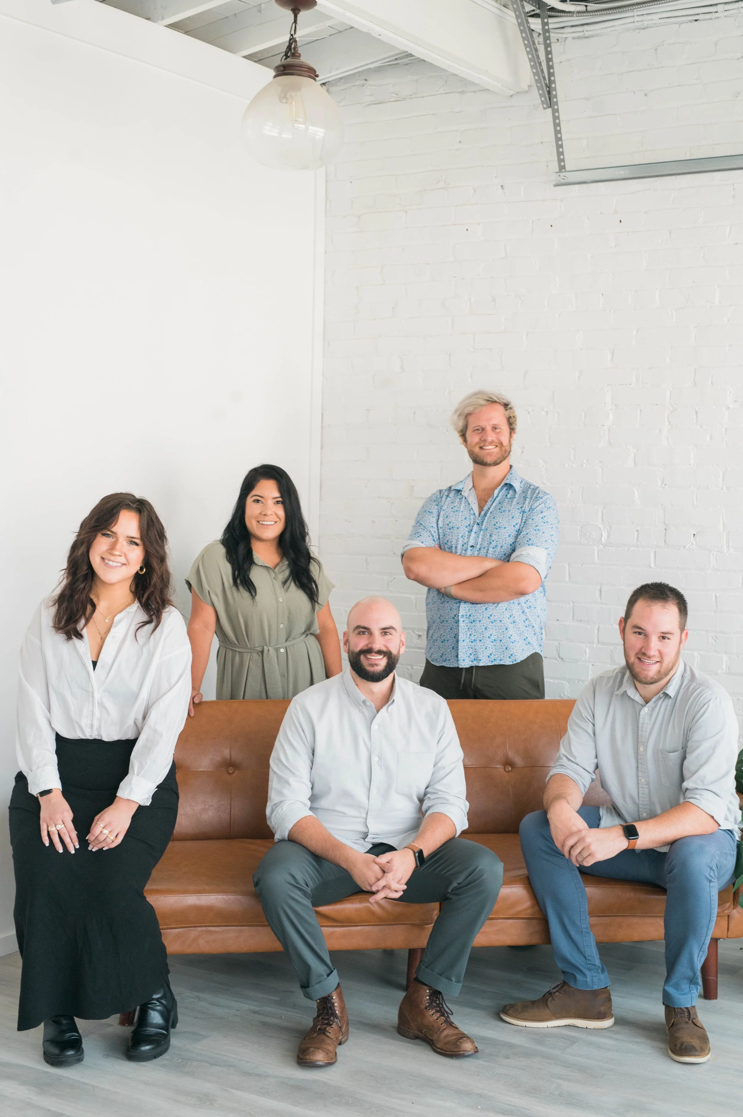 Five people posing together, with three seated on a brown leather sofa and two standing behind them, in a bright room with white brick walls.