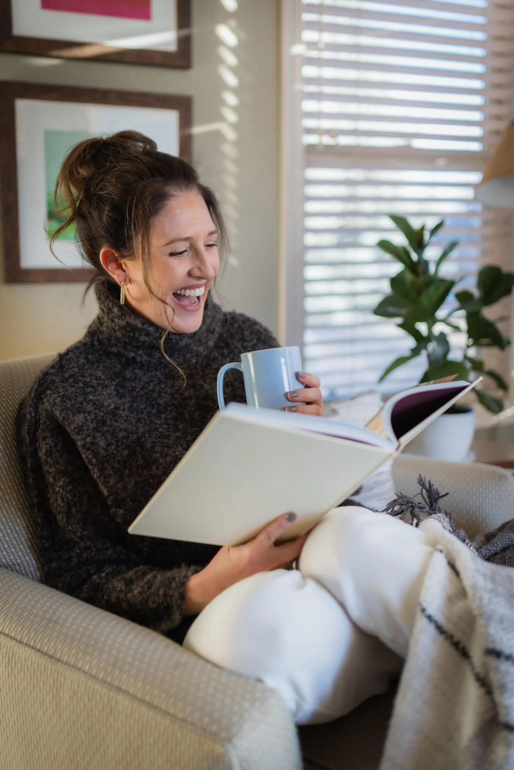 A woman sitting on a chair, reading a book and holding a blue mug, smiling. She is wearing a dark knitted sweater and has her hair tied up. The room has framed pictures on the wall and a potted plant is visible. Sunlight is streaming through blinds.