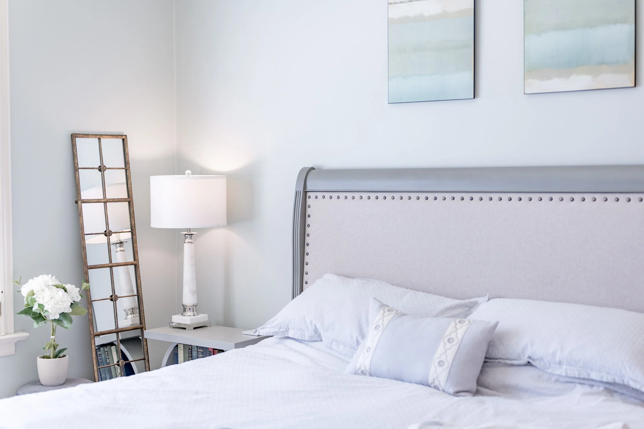 A cozy bedroom with a light blue wall features a neatly made bed with soft pillows and a decorative headboard. A white bedside table holds a white lamp. A leaning mirror reflects the room, and a vase with white flowers sits next to it. Two abstract p